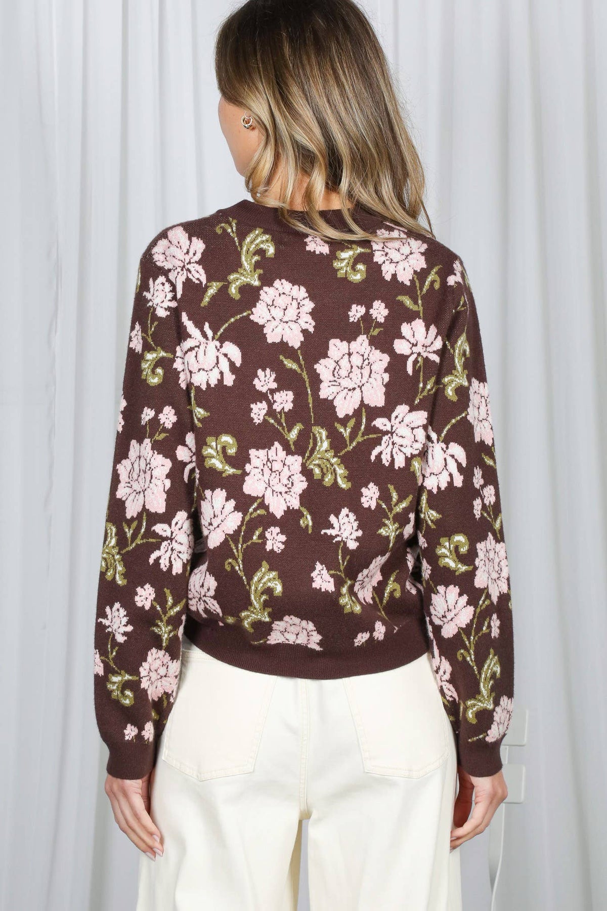 A woman viewed from the back wearing a brown long-sleeve knit sweater with a pink and green floral pattern, paired with cream-colored pants, set against a white curtain background.