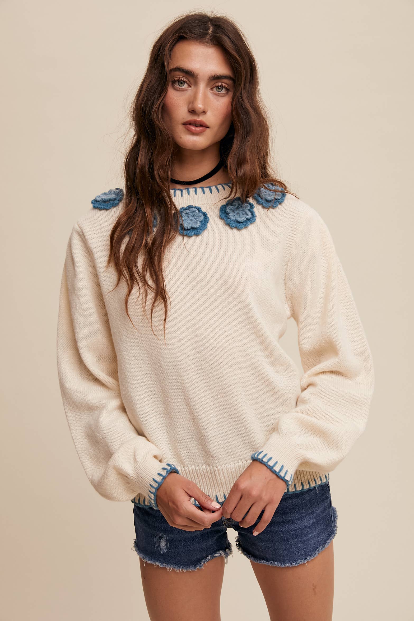 Cream sweater with blue crochet flower appliqués at neckline, blue whipstitch trim at cuffs and hem, long sleeves; styled with frayed denim shorts.