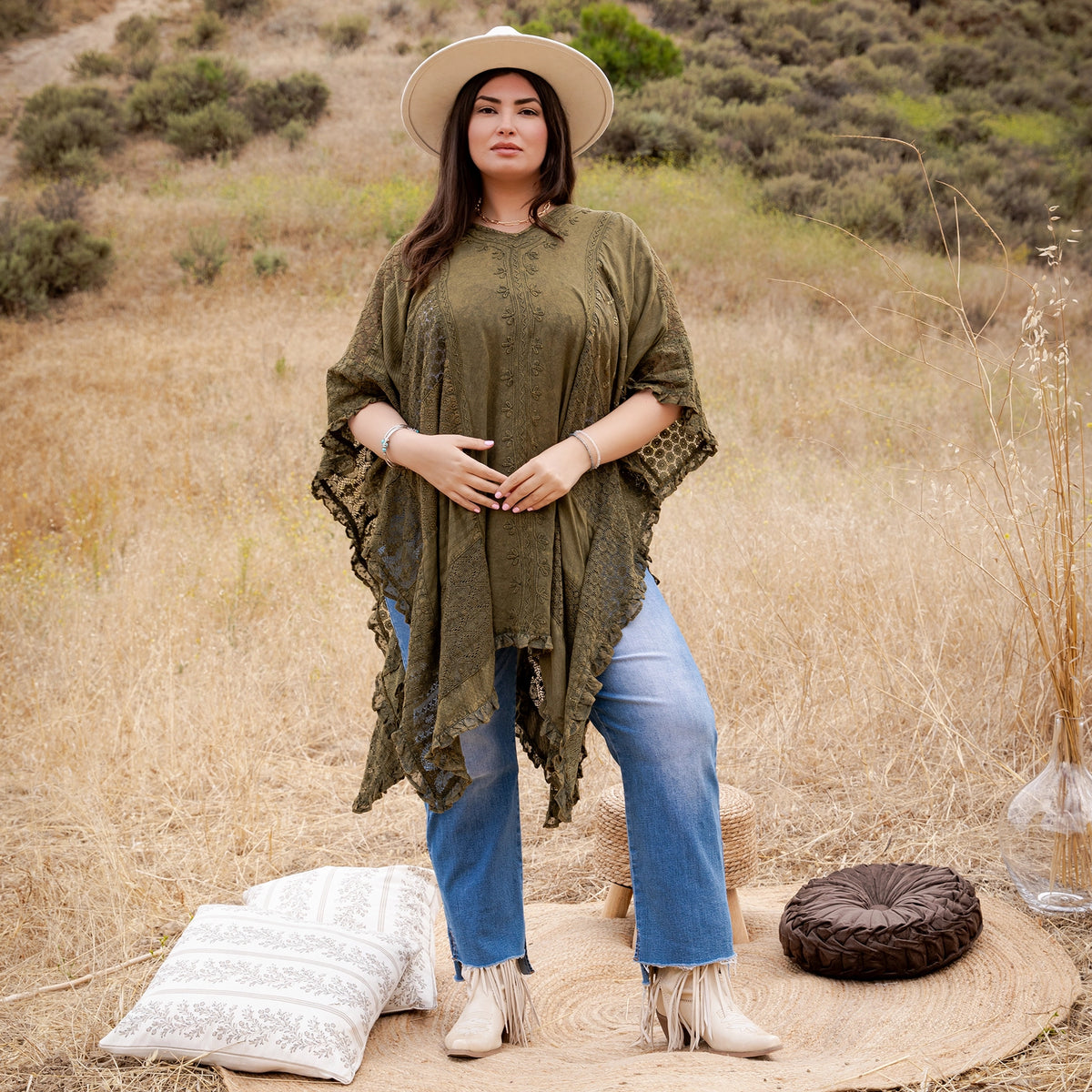 A woman stands outdoors in a dry grassy field wearing an olive green lace poncho, blue jeans, and white fringe boots, with a straw hat. A woven rug, decorative pillow, and small round cushion are on the ground beside her.