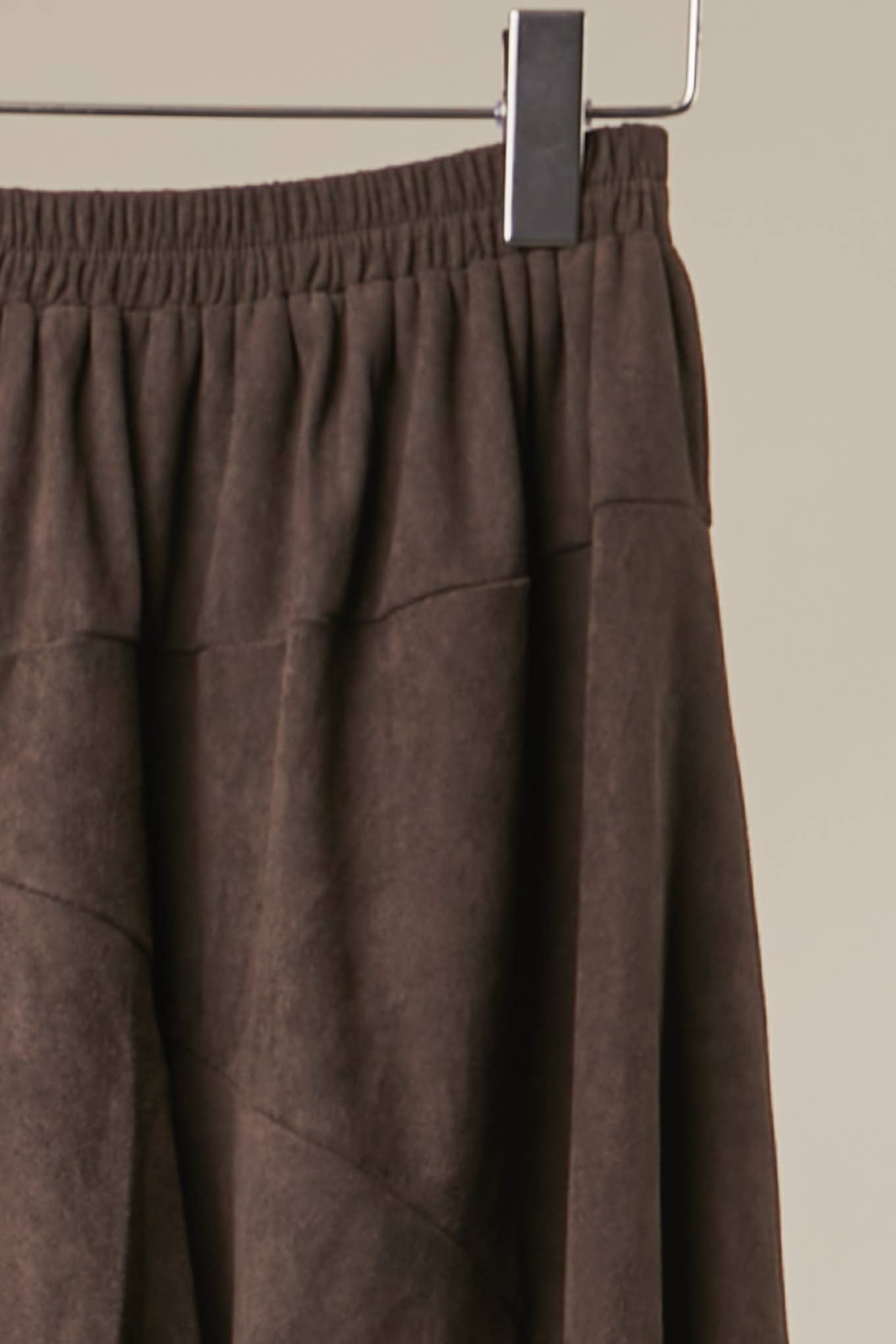 A close-up of a dark brown textured skirt with an elastic waistband, hanging from a metal hanger against a solid beige background.