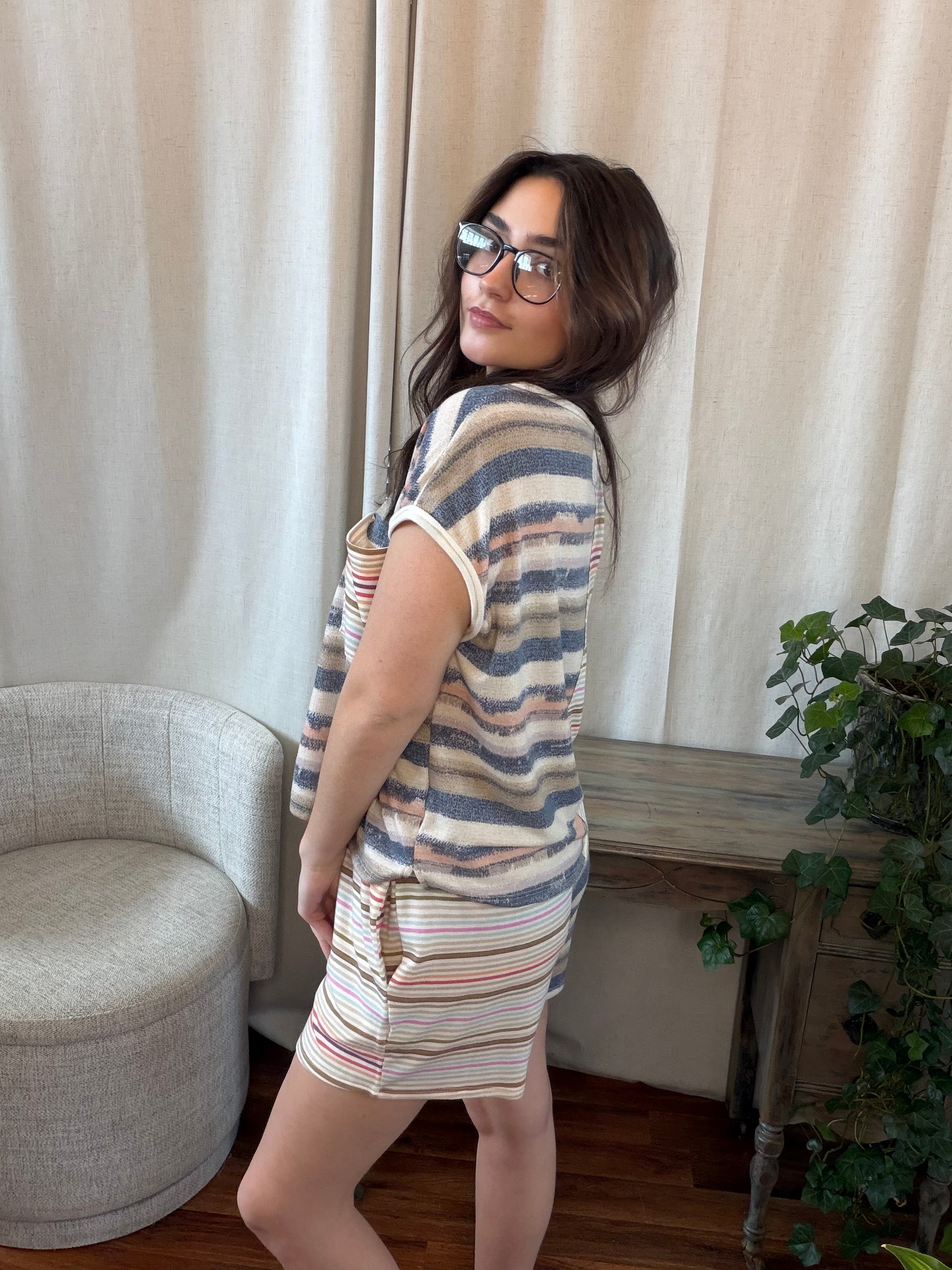Woman wearing a striped outfit standing in a room with curtains and a plant.