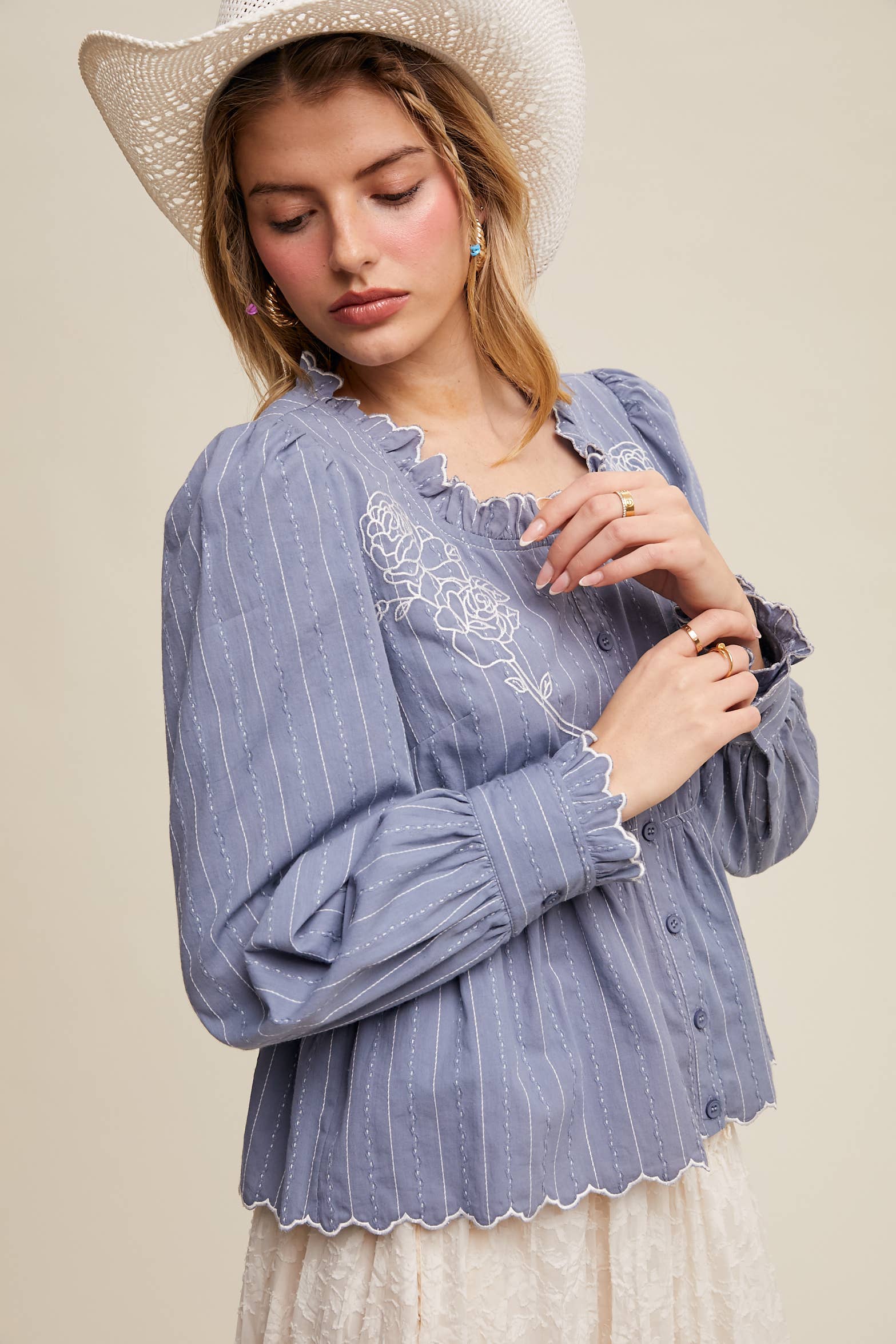 Side view- Dusty blue pinstripe blouse with ruffled neckline, white rose embroidery, puff long sleeves, button front, and scalloped peplum hem—styled with a white lace skirt and cowboy hat.