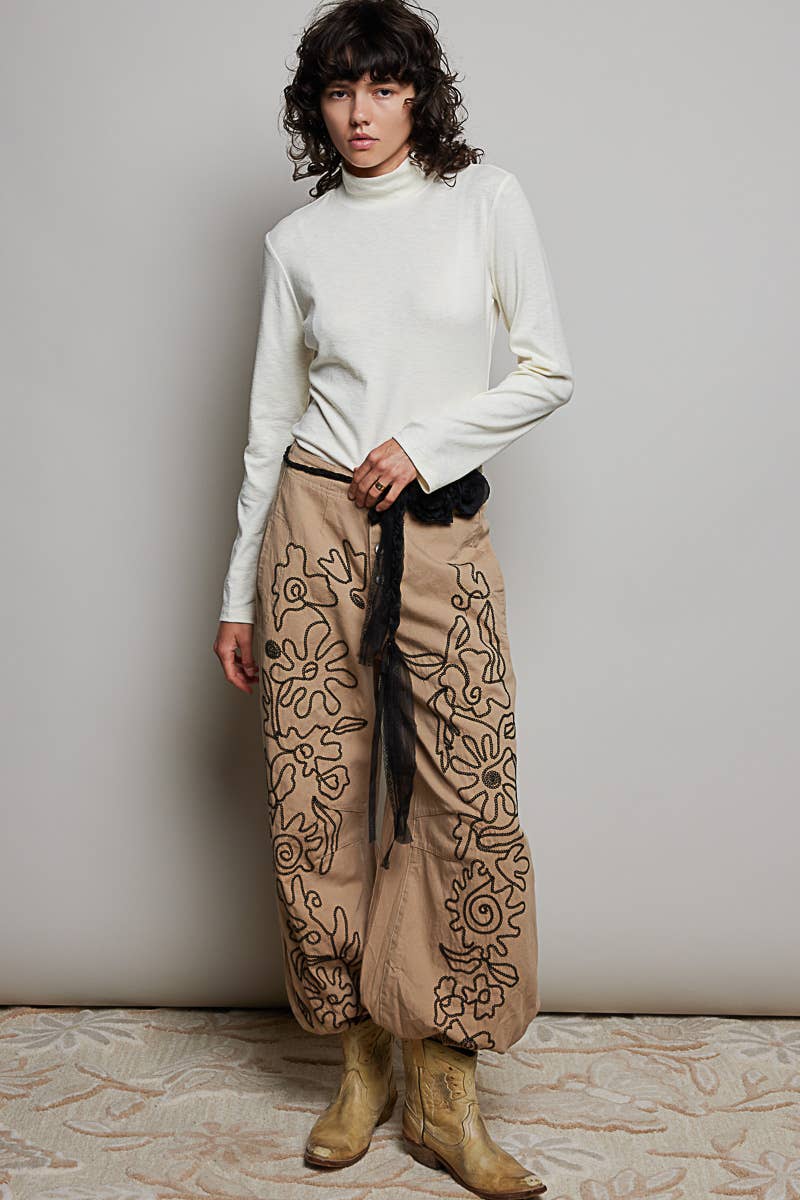 Person wearing a white turtleneck and patterned beige pants against a plain background
