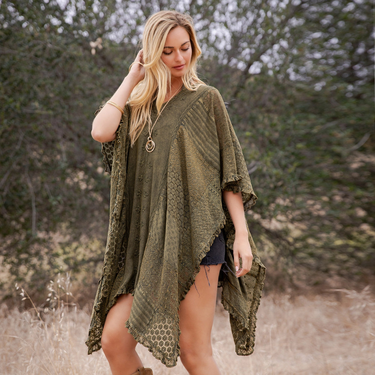 A woman stands outdoors in a grassy field with trees in the background, wearing an olive green bohemian lace poncho top over dark denim shorts.