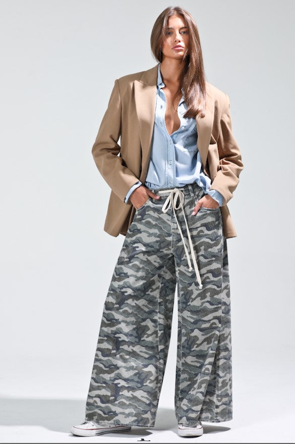 Woman wearing a beige blazer, light blue shirt, and camouflage pants on a white background