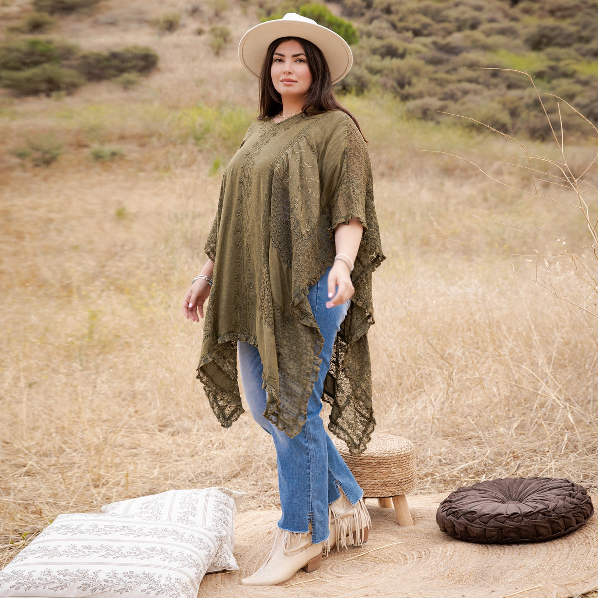 A woman stands outdoors in a dry grassy field wearing an olive green lace-trim poncho top, blue jeans, and beige boots, with a white hat. A woven stool, patterned pillow, and round woven cushion are on the ground beside her.