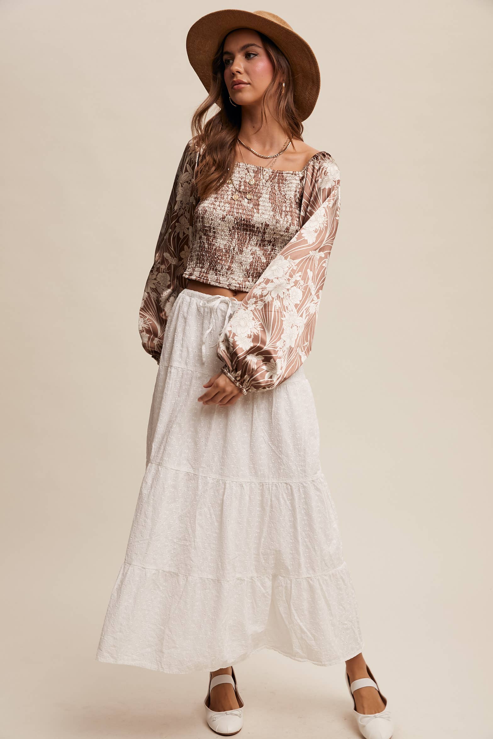 Brown and cream floral smocked blouse with square neckline, puffed long sleeves, and elastic cuffs, styled with layered necklaces and a wide-brim hat.