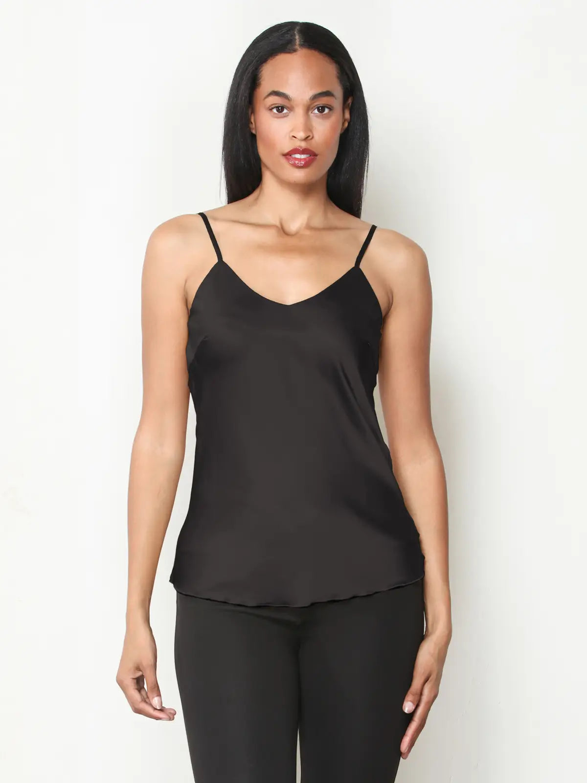 A woman modeling a black satin camisole top with thin spaghetti straps and black leggings against a plain white studio background.
