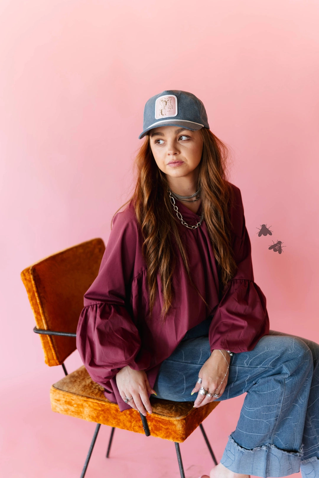 A young woman with long brown hair sits on an orange velvet chair against a solid pink studio background. She is wearing a dark gray baseball cap with a white graphic, a dark burgundy long-sleeve blouse with dramatic puffed sleeves, and blue jeans.