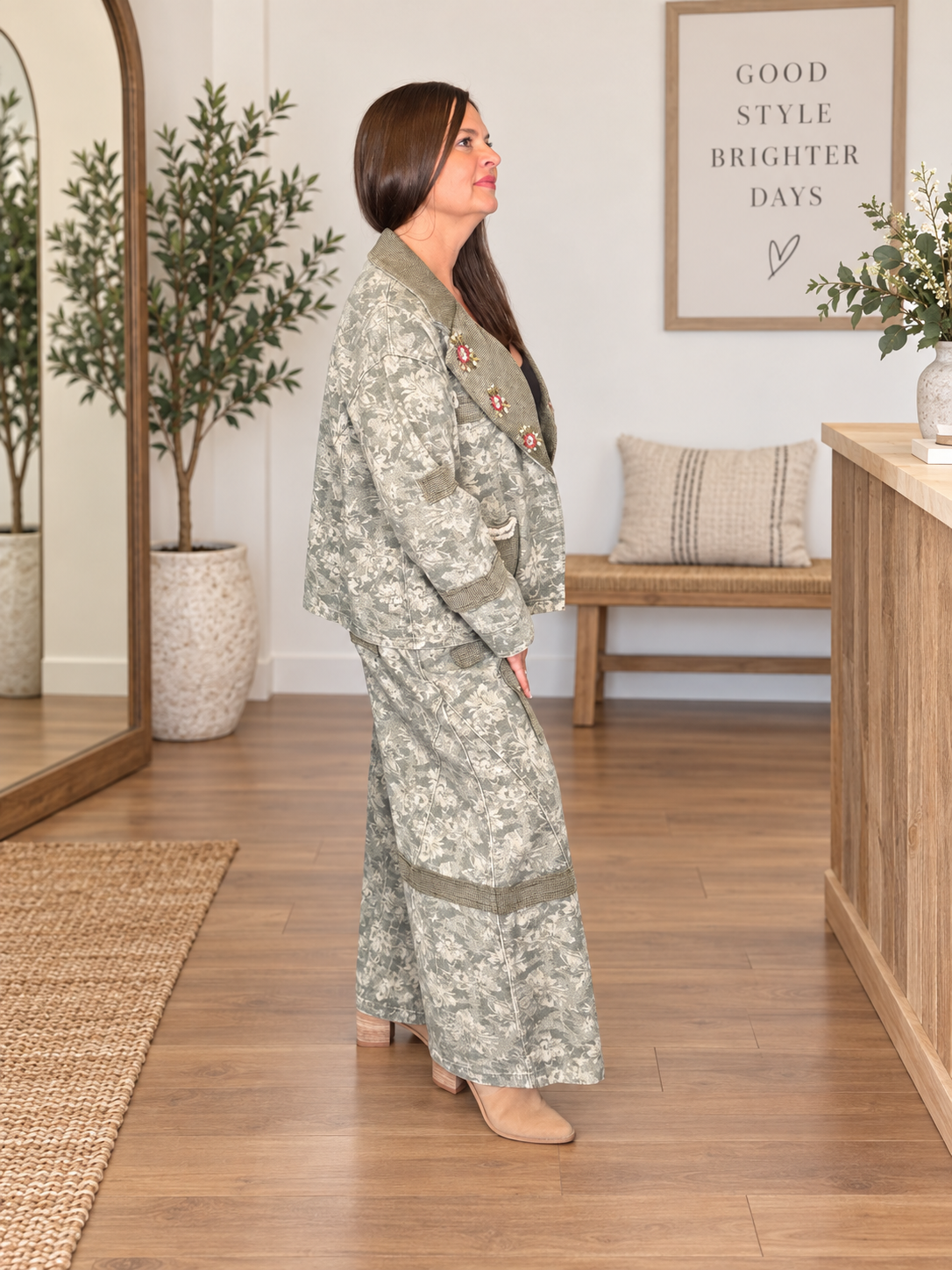 A woman wearing a sage green and white floral print robe with olive green trim, standing in a bright, modern room with wood floors and a wooden bench in the background.