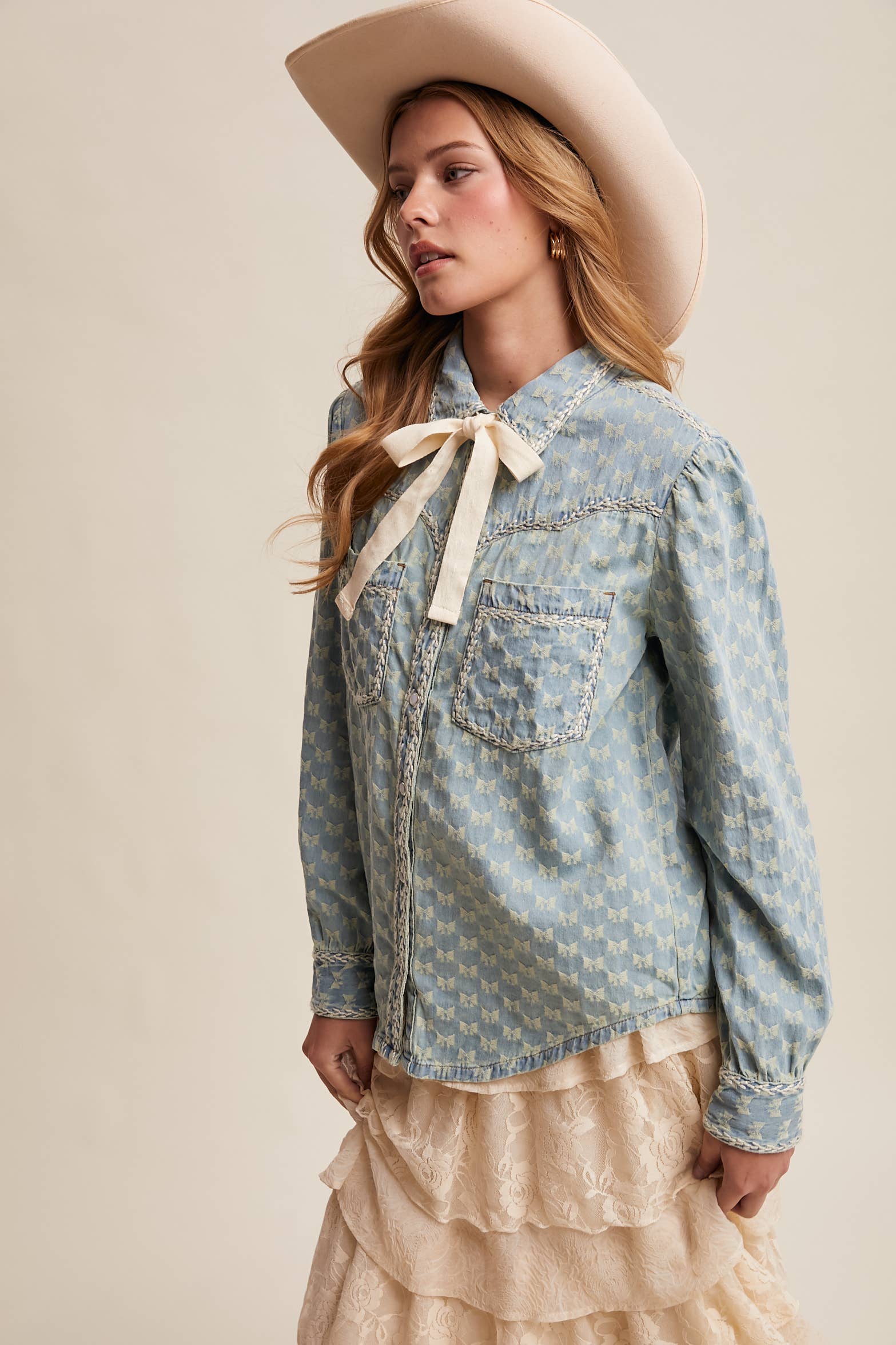Side view- Light-wash denim blouse with cream bow-tie collar, tonal mini-bow pattern, braided contrast stitching, snap front, and two chest pockets.