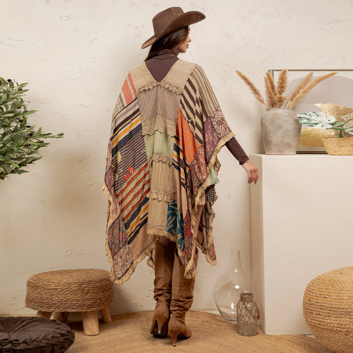 A woman stands in profile wearing a brown wide-brim hat and a long patchwork poncho with fringe, paired with brown boots, in a styled indoor room with woven stools, a plant, and a white display block.