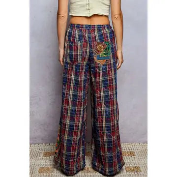 A person wearing high-waisted, wide-leg plaid pants with a colorful embroidered patch on the back pocket, shown from the waist down against a textured gray wall.