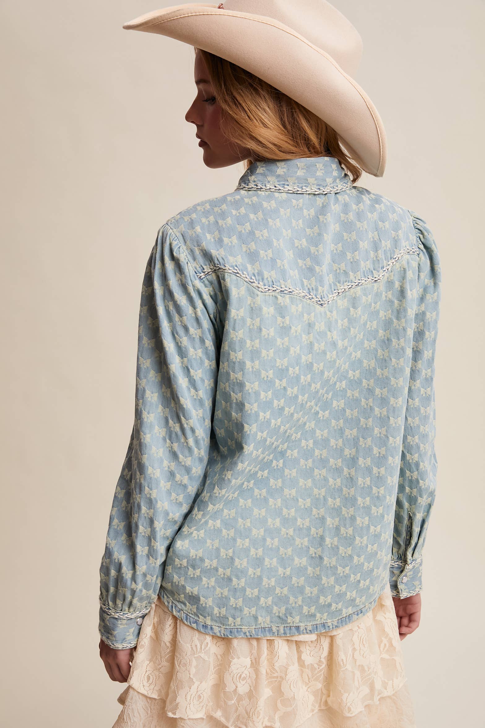 Back view- Light wash denim blouse with cream bow-tie collar, tonal mini-bow pattern, braided contrast stitching, snap front, and two chest pockets.