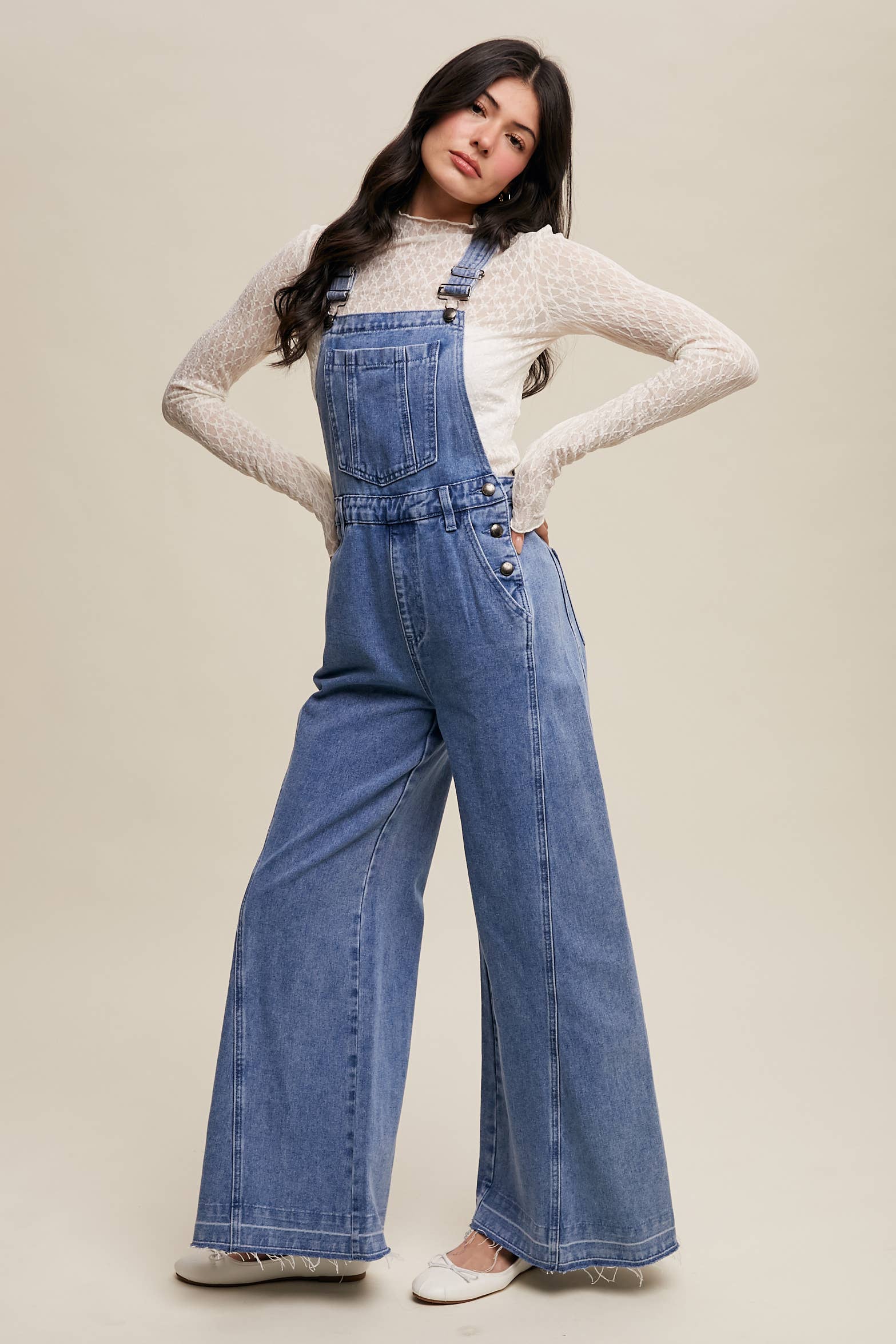 Side view- Light-wash wide-leg denim overalls with adjustable straps, front bib pocket, side buttons, and slant pockets, finished with a raw hem and layered over a cream long-sleeve top.
