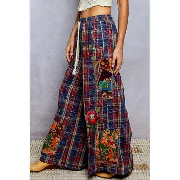 A person wearing wide-leg plaid pants with colorful floral and geometric patchwork details, paired with tan boots against a gray studio backdrop.