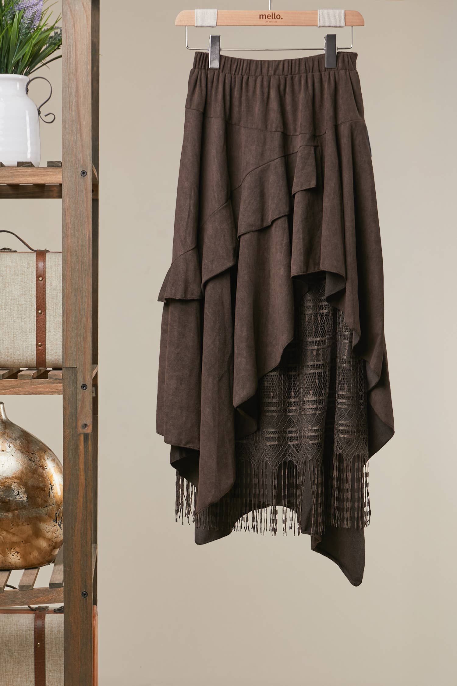 A dark brown tiered asymmetrical skirt with fringe detailing, hanging on a wooden hanger against a beige wall.