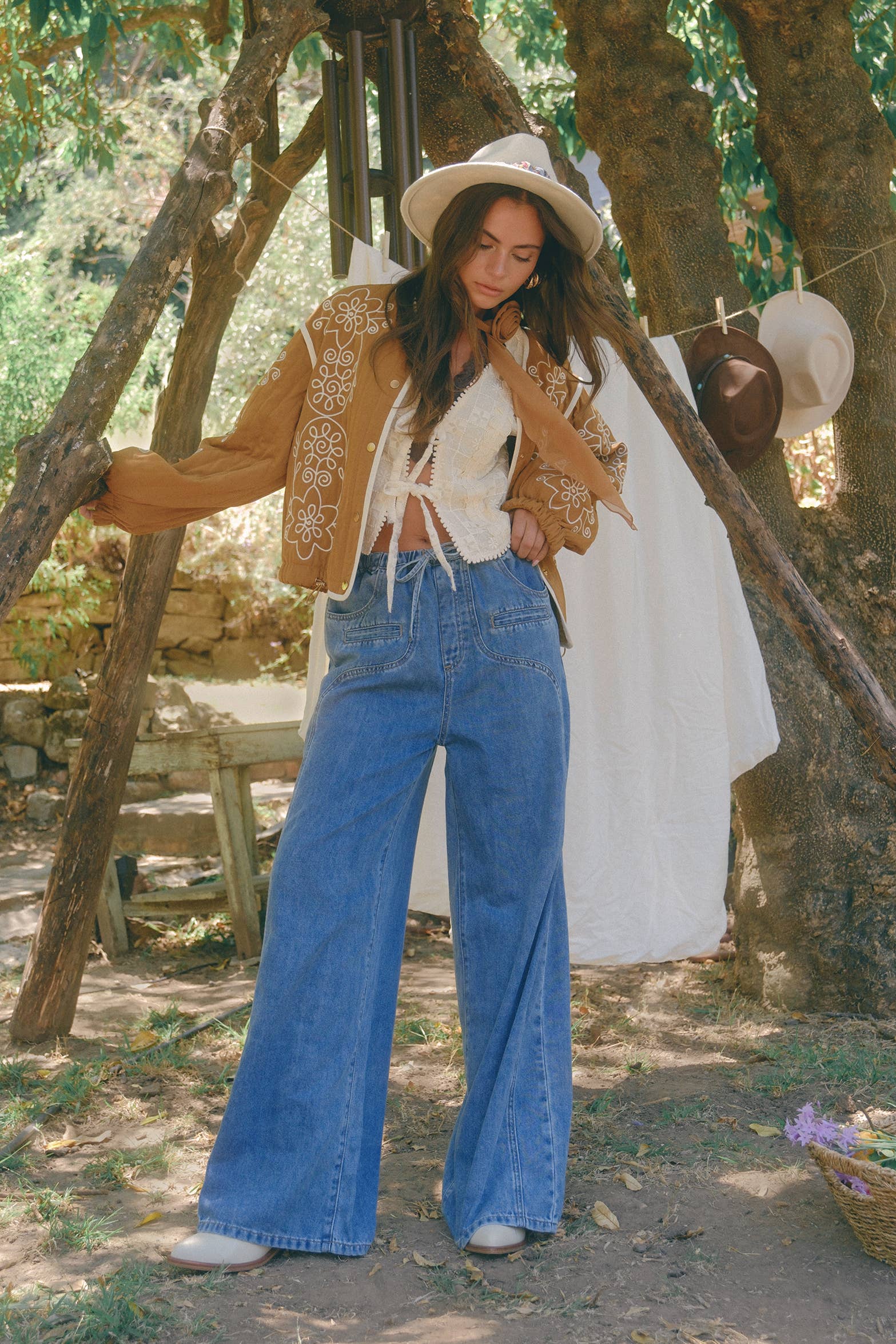 Boho caramel-brown jacket with cream floral swirl embroidery, ivory piping, and snap buttons, styled over a lace tie-front top with a scarf and felt hat outdoors.
