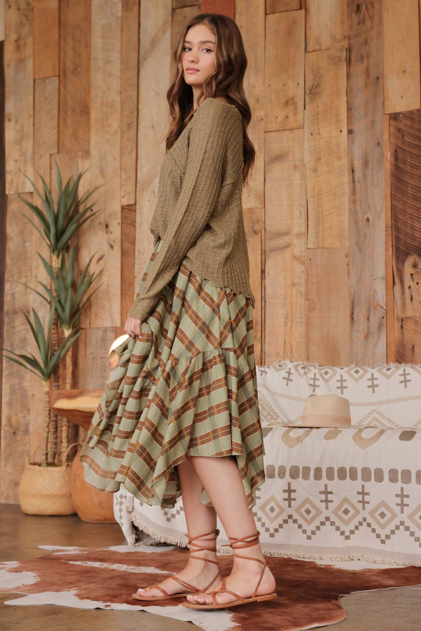 Side view of model wearing olive distressed lightweight sweater with ribbed knit texture and raw edge hem, paired with a green and brown plaid ruffle skirt and lace-up sandals.