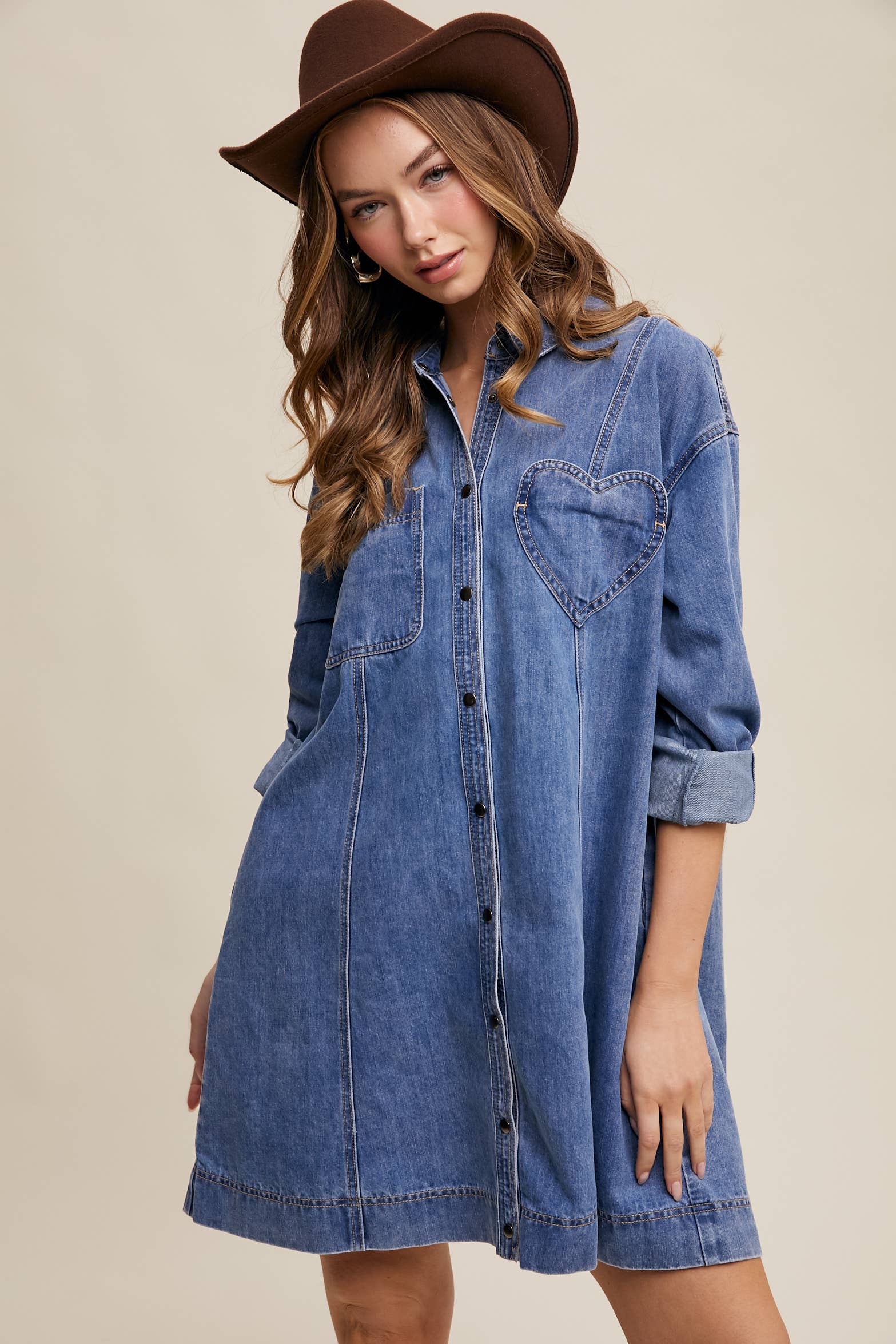 Medium-wash denim shirt dress with snap button front, heart-shaped chest pocket, rolled long sleeves, and a relaxed mini silhouette.