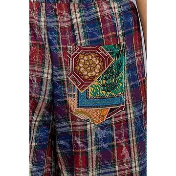 Close-up of a plaid garment, likely shorts or pants, featuring a multicolor patchwork pocket with geometric and floral patterns over red, blue, and green plaid fabric.