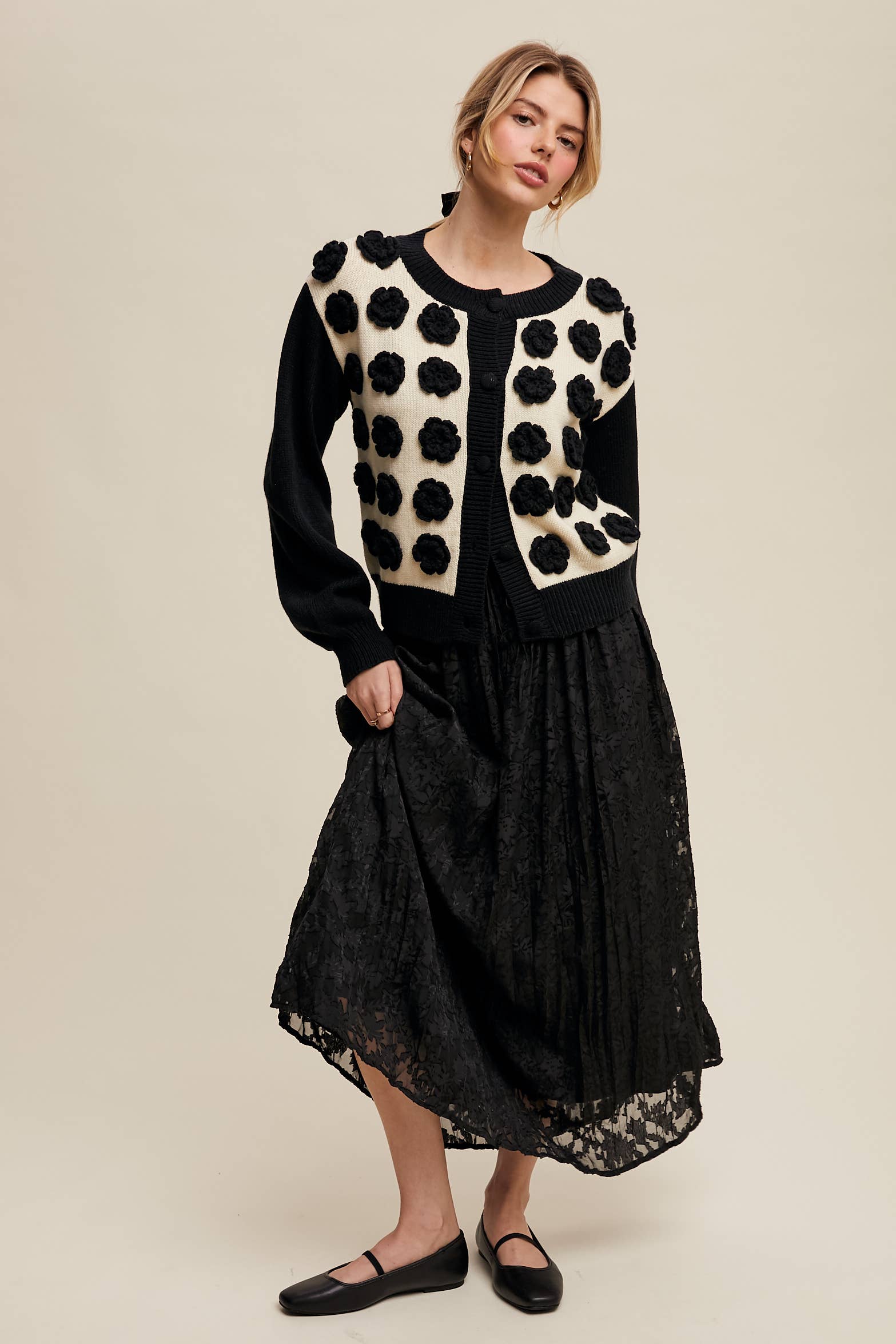 Black floral lace maxi skirt with drawstring waist and subtle bubble hem, styled with a cream-and-black 3D flower cardigan and black flats.