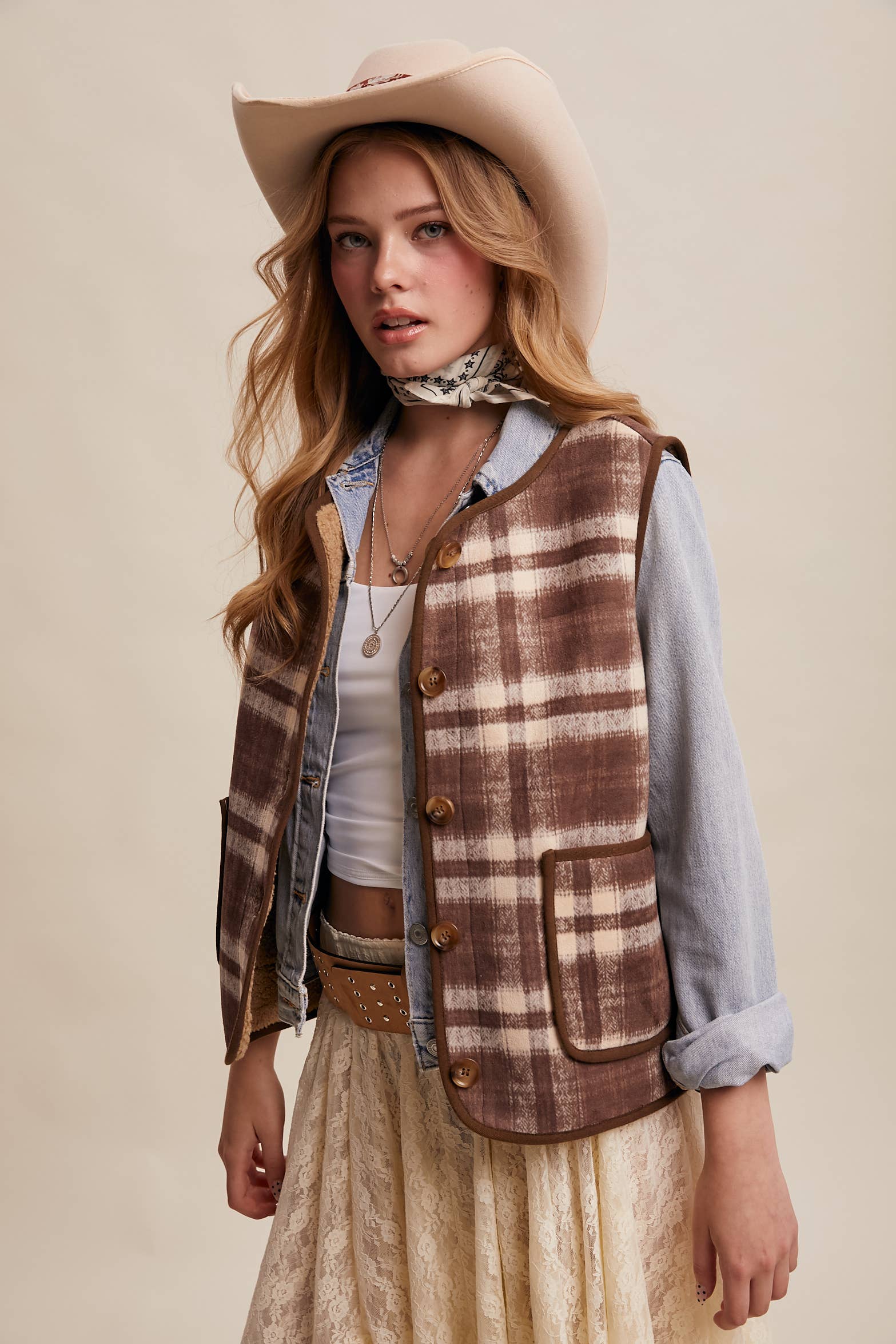 Woman wearing a plaid vest, cowboy hat, and scarf on a beige background