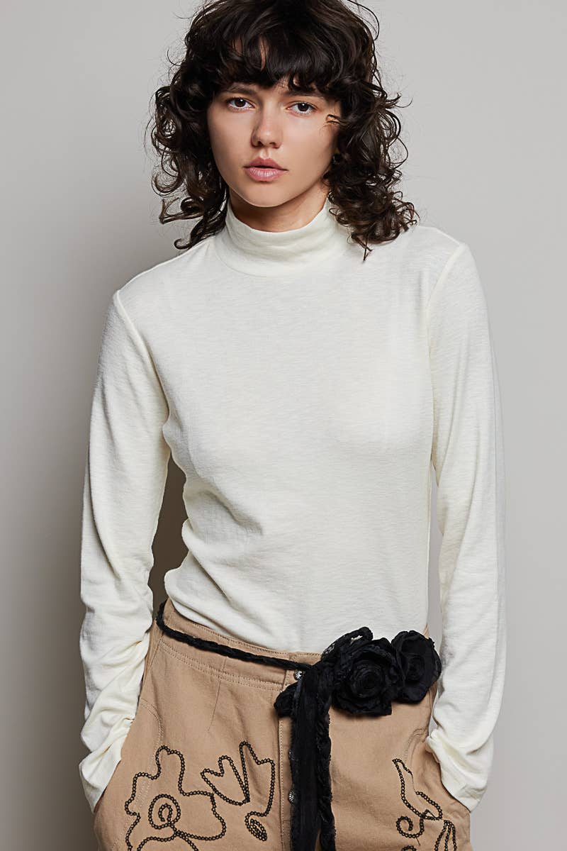 A model with dark curly hair wearing a cream-colored mock neck long-sleeve top and beige pants with a black fabric flower belt tie, photographed against a neutral gray background.