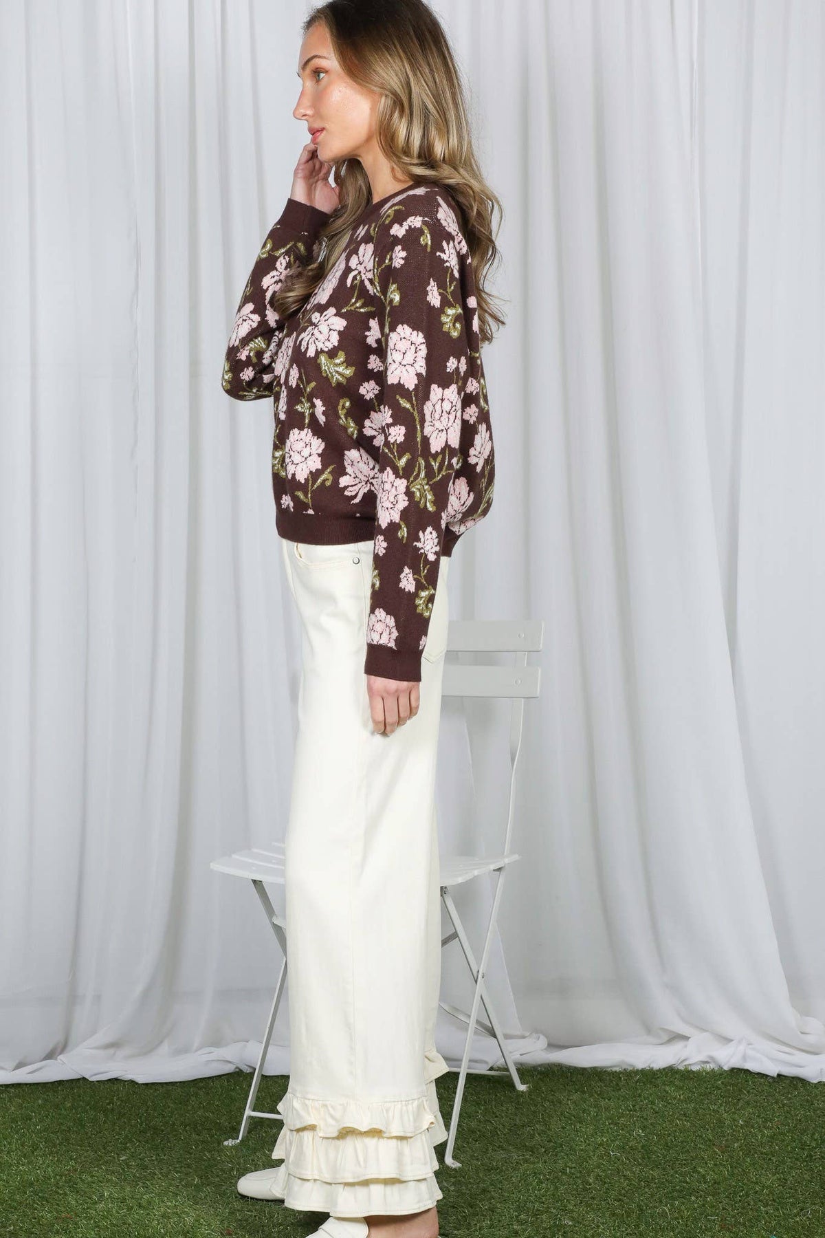 A woman stands in profile wearing a brown floral long-sleeve top and cream ruffled wide-leg pants, posed in front of a white draped backdrop with artificial green turf at the bottom and a white folding chair behind her.