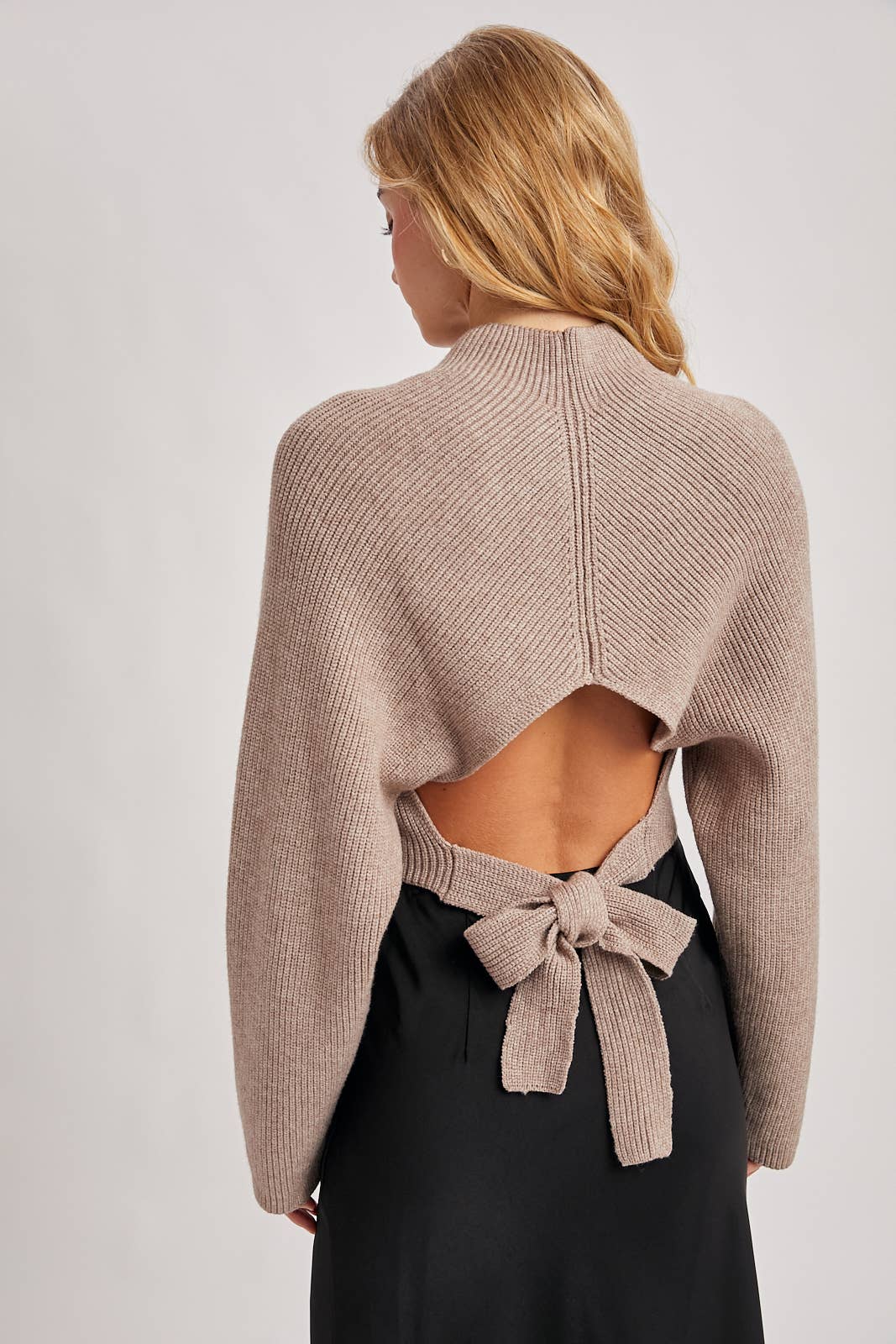 Back view of taupe ribbed open-back sweater with long sleeves and a waist tie bow, styled with a black skirt.