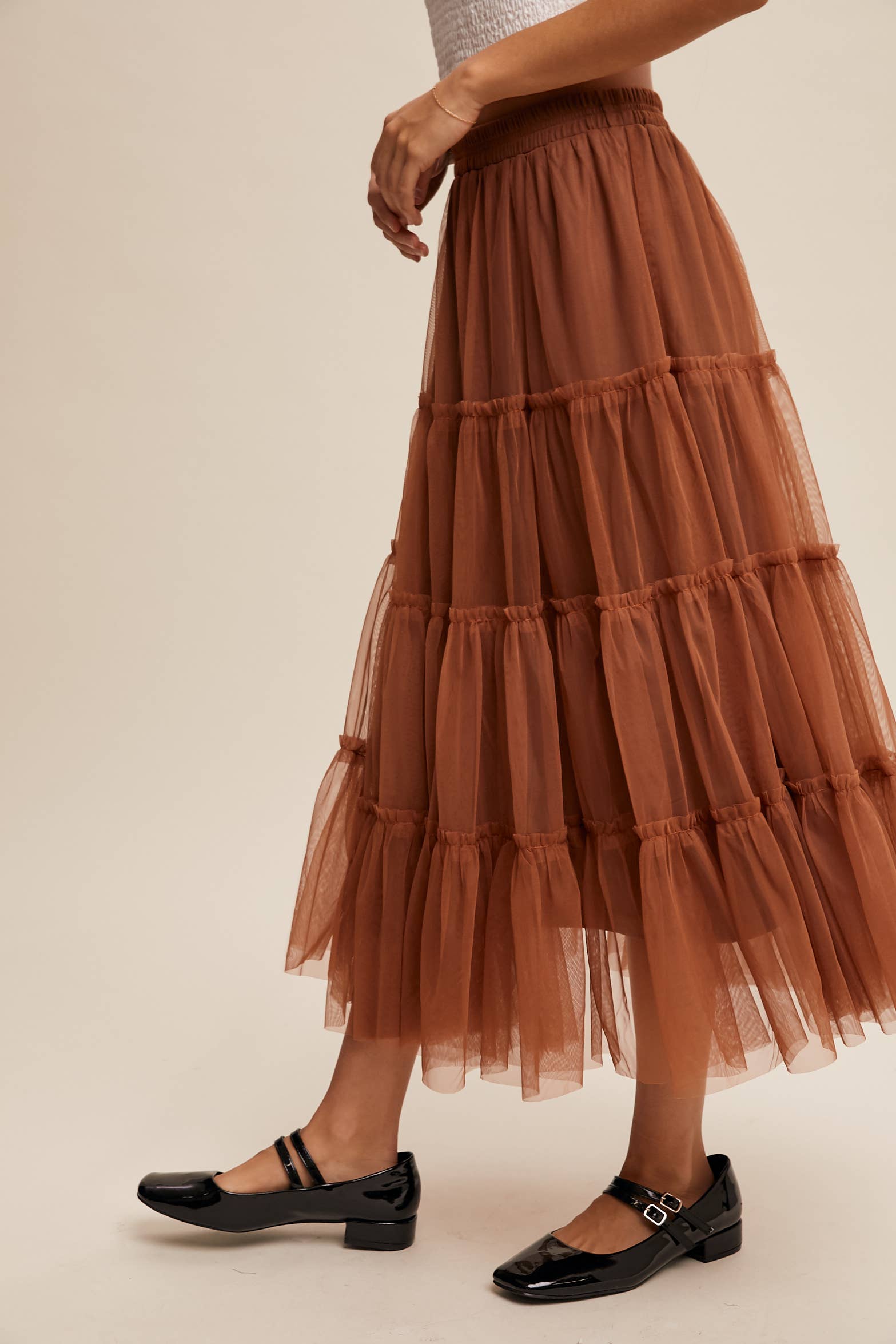 Side view — rust tulle tiered midi skirt with elastic waist and ruffle tiers, sheer overlay; model in black Mary Jane flats.