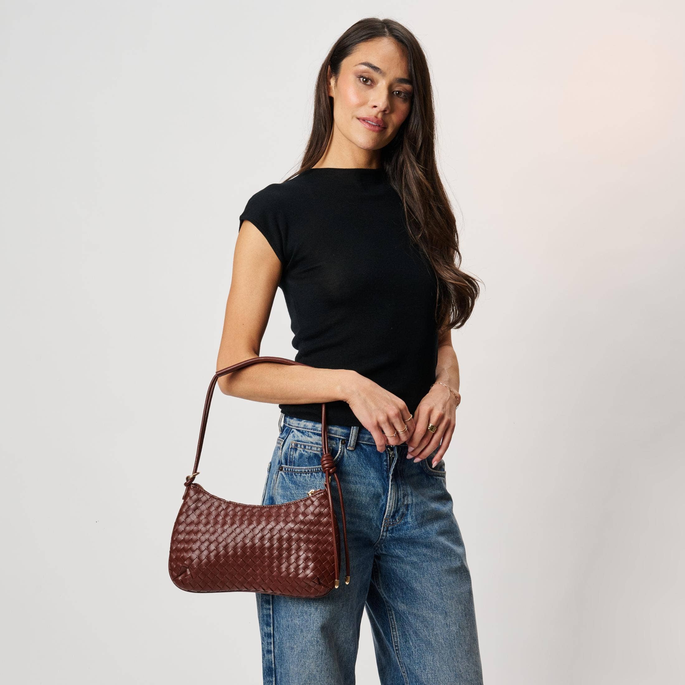 Cindy Woven Crescent Shoulder Bag in Burgundy by Moda Luxe — close-up model carry view showing woven vegan leather crescent silhouette, knotted strap detail, and gold-tone zip pull, styled with black top and denim jeans