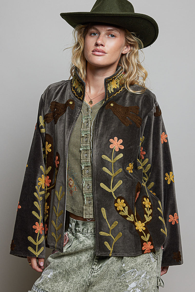 A woman wearing a dark brown velvet jacket with colorful floral embroidery, an olive green wide-brim hat, and distressed green jeans, photographed against a neutral gray studio background.