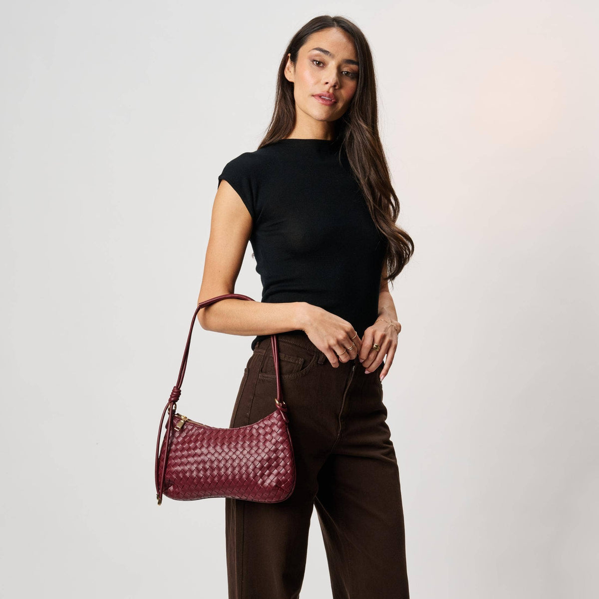 Cindy Woven Crescent Shoulder Bag in Burgundy by Moda Luxe — model shoulder carry view showing woven vegan leather crescent bag with knotted strap, gold-tone hardware and zip closure, styled with black top and dark brown trousers
