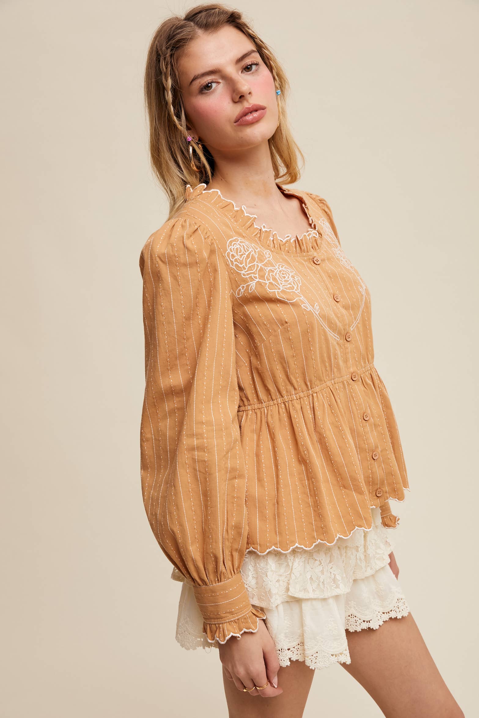 Side view- Camel pinstripe blouse with ruffled neckline, white rose embroidery, puff long sleeves, button front, and scalloped peplum hem—styled with white lace shorts.