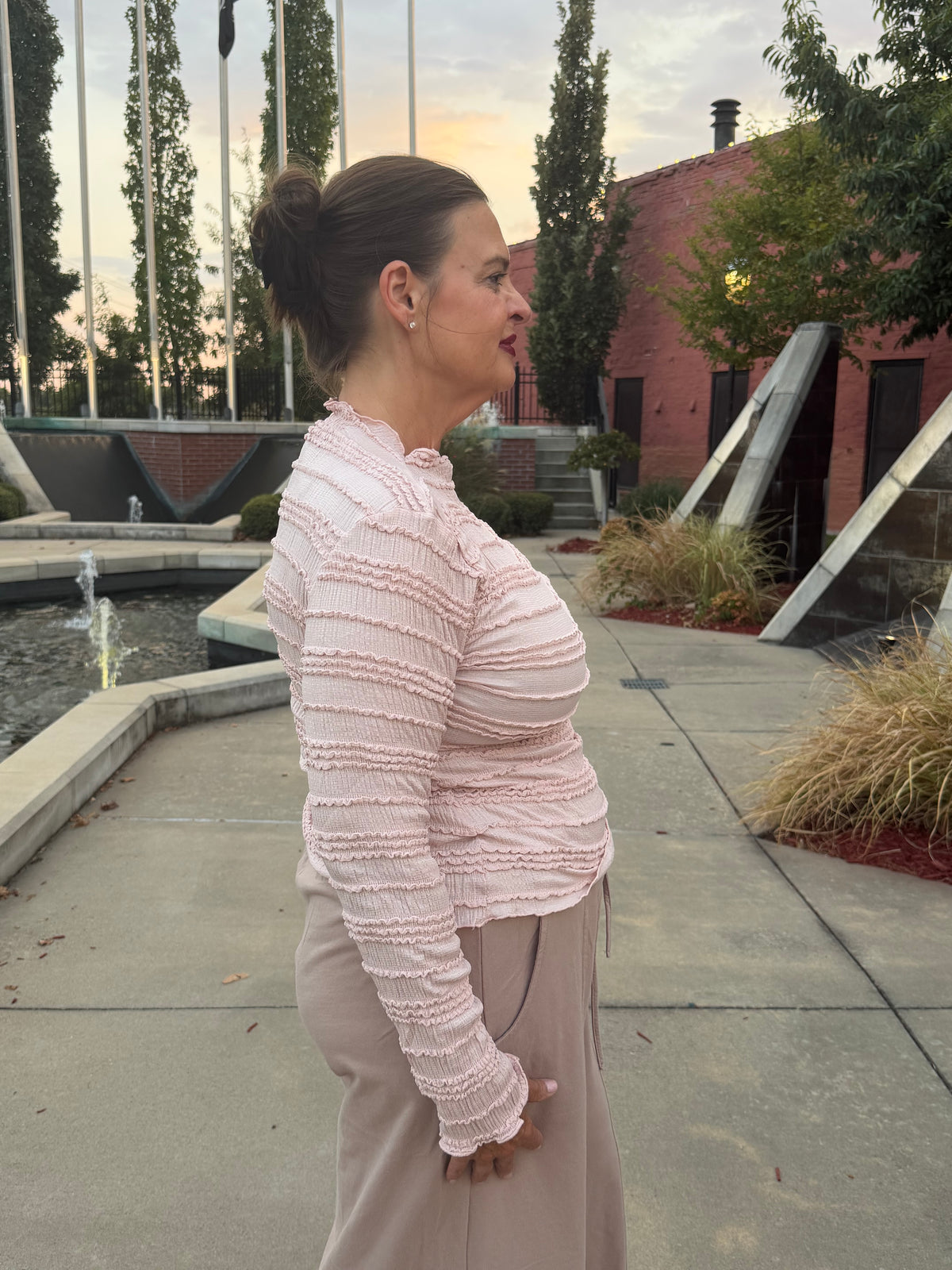 A woman wearing a light pink textured long-sleeve top and beige pants, standing outdoors in a landscaped area with a fountain, trees, and a brick building in the background.