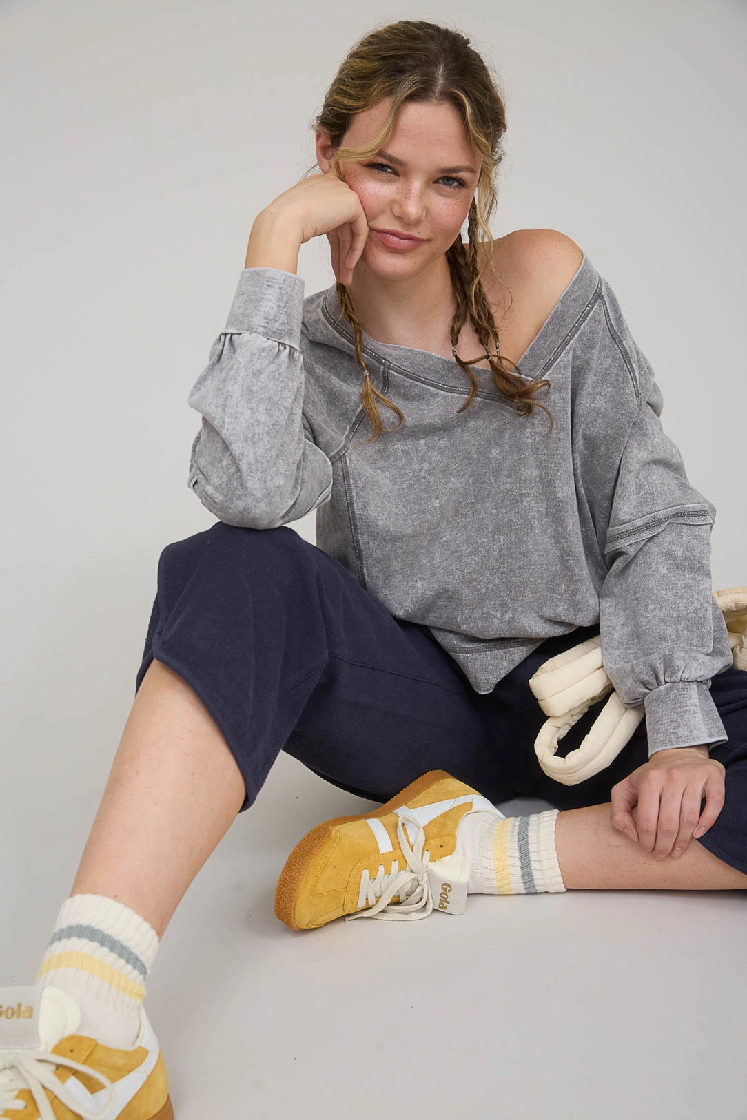 Woman wearing a gray off-shoulder sweater and navy pants, sitting on a light-colored floor.