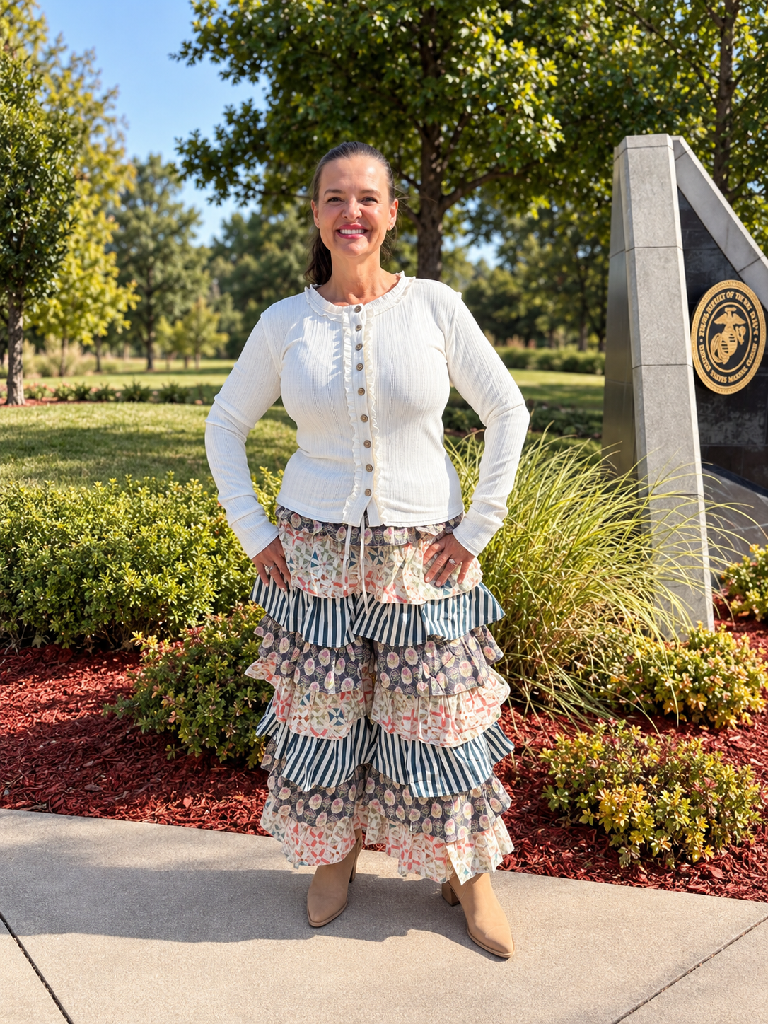 A woman stands outdoors in front of trees and landscaping, wearing a white long-sleeve top and a long tiered ruffled pants from bluevelvet with striped and floral patterns.