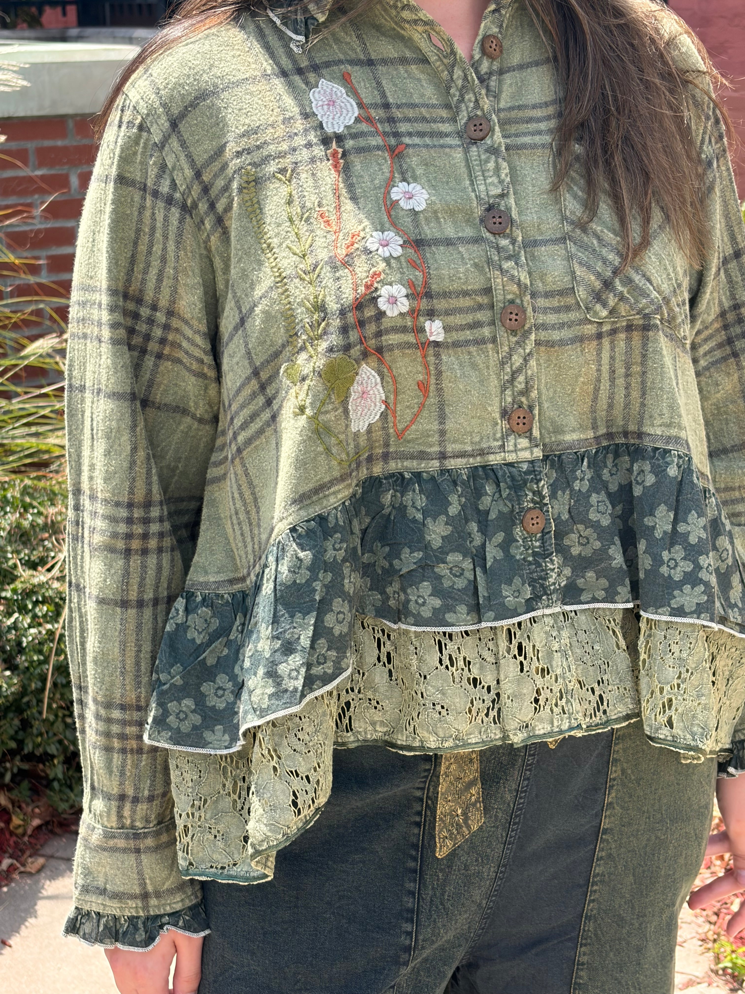A person wearing a patchwork-style long-sleeve blouse with a muted green plaid upper section, embroidered floral detail, a dark teal floral ruffle band, and a cream lace ruffle hem, paired with dark jeans in an outdoor setting.