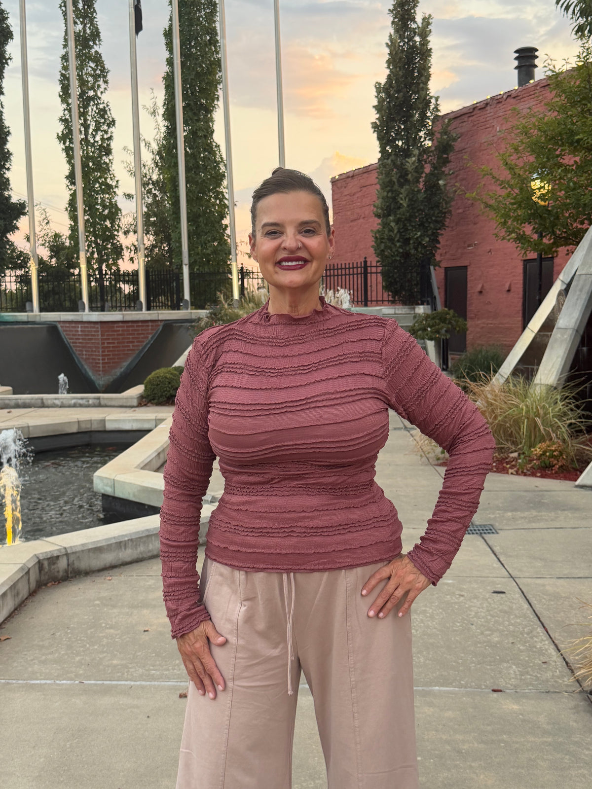 A woman wearing a ribbed, long-sleeve mock neck top in dusty rose and beige drawstring pants, standing outdoors in front of a brick building and fountain.