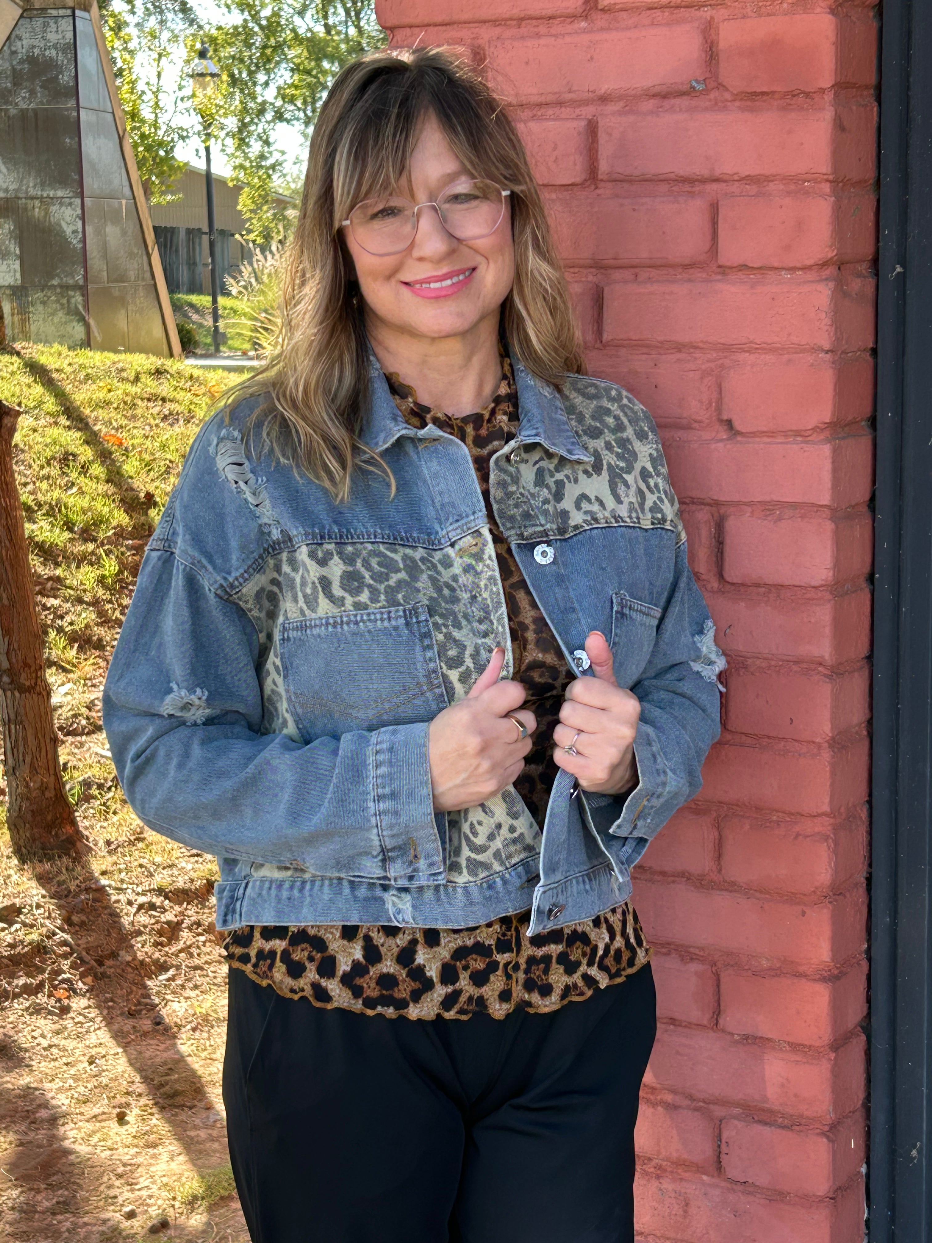Person wearing a denim jacket with leopard print accents, standing against a brick wall.