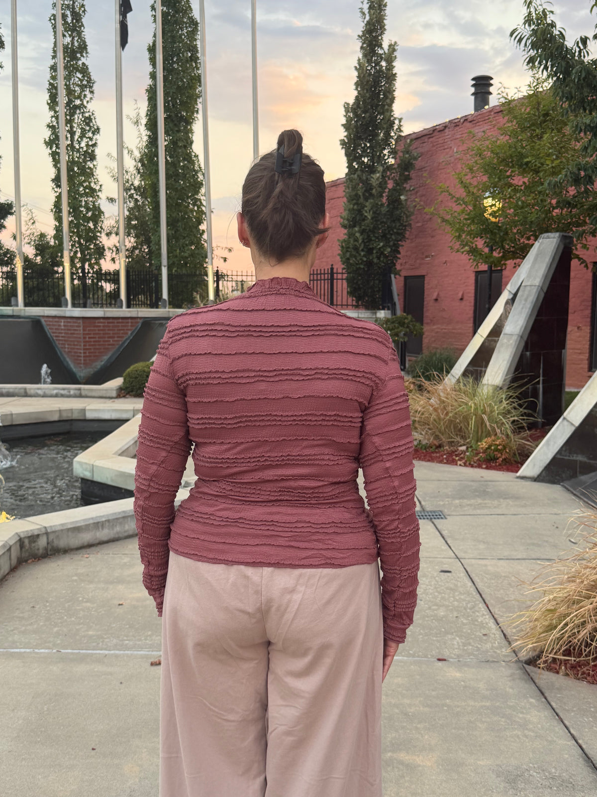 A woman viewed from behind wearing a dusty rose ribbed long-sleeve mock-neck top and light pink wide-leg pants, standing outdoors in a landscaped courtyard with a brick building, flagpoles, and a concrete walkway.