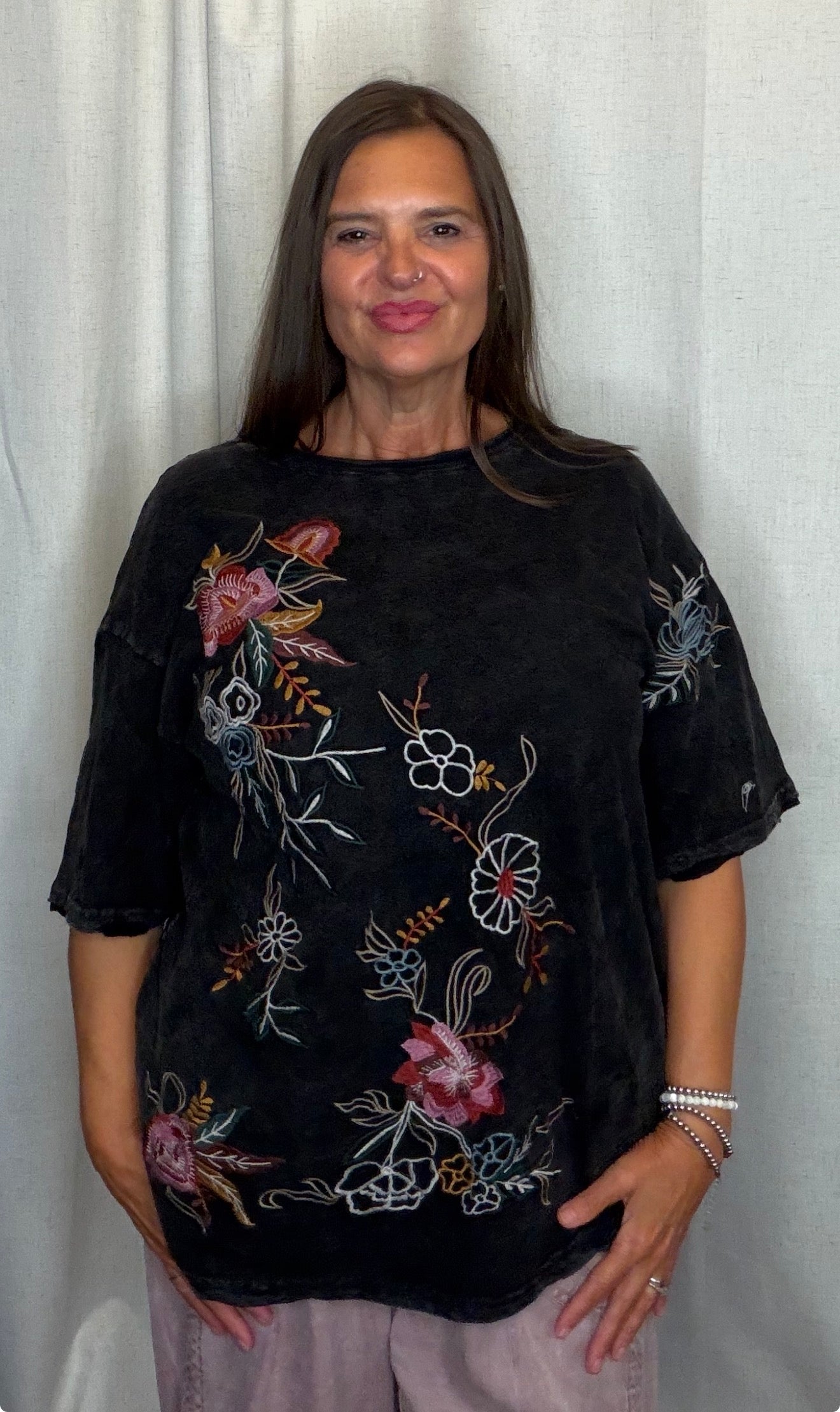 A woman modeling a black short-sleeve t-shirt with colorful floral embroidery, paired with light pink pants, standing against a white curtain backdrop.