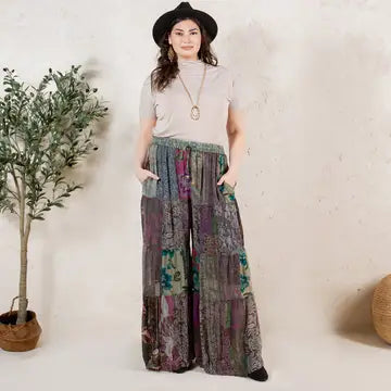 Woman wearing a white t-shirt and patterned wide-leg pants standing against a plain wall with a plant to the left.