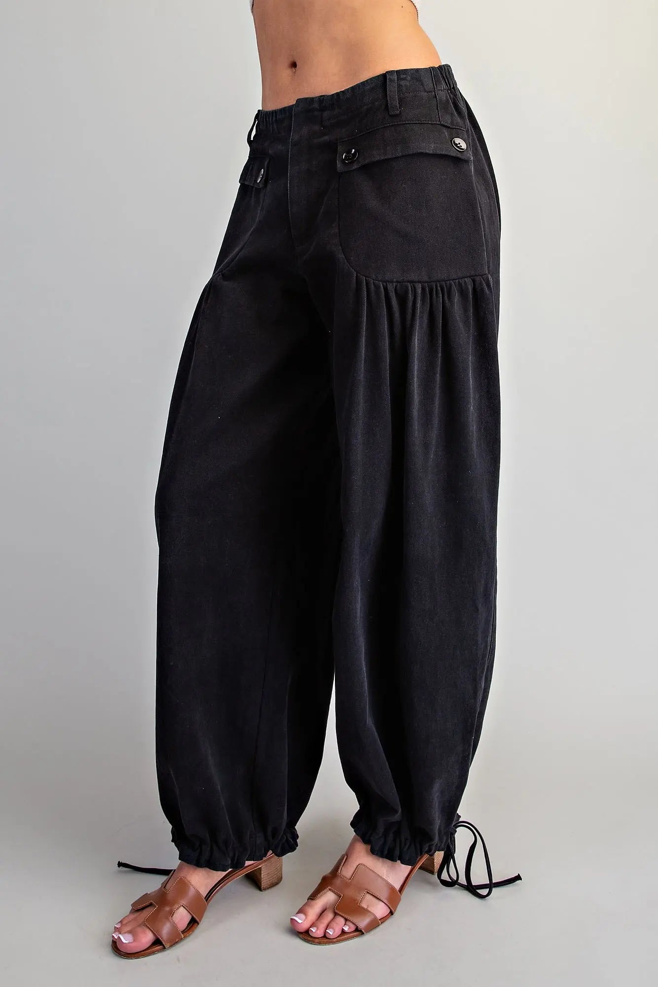 Black wide-leg harem pants with gathered balloon legs, elastic cuffs, and side tie details, shown on a model wearing brown heeled sandals against a light gray studio background.