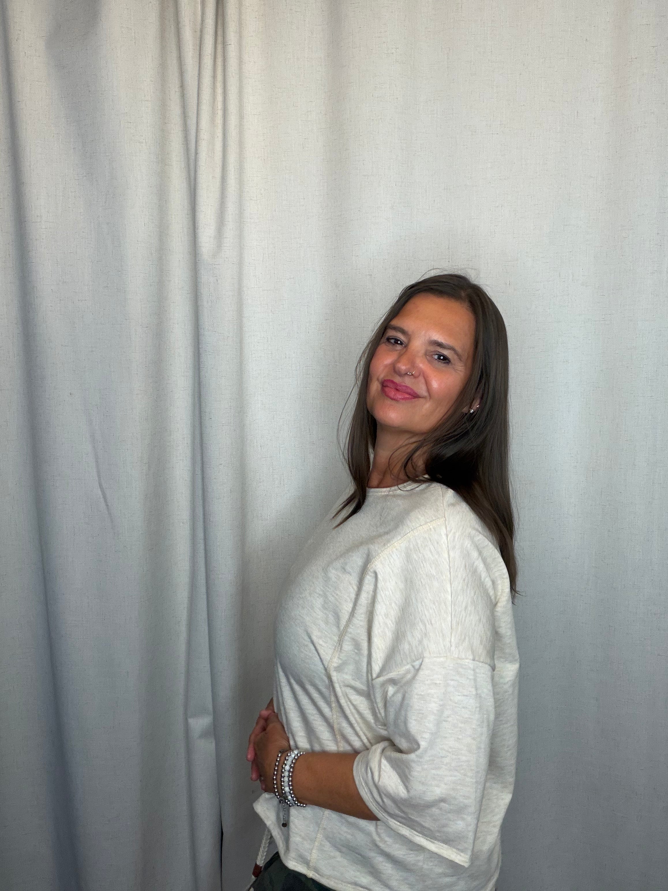 A woman with long brown hair is shown from the side, wearing a light beige textured long-sleeve top, standing in front of a light gray fabric backdrop.