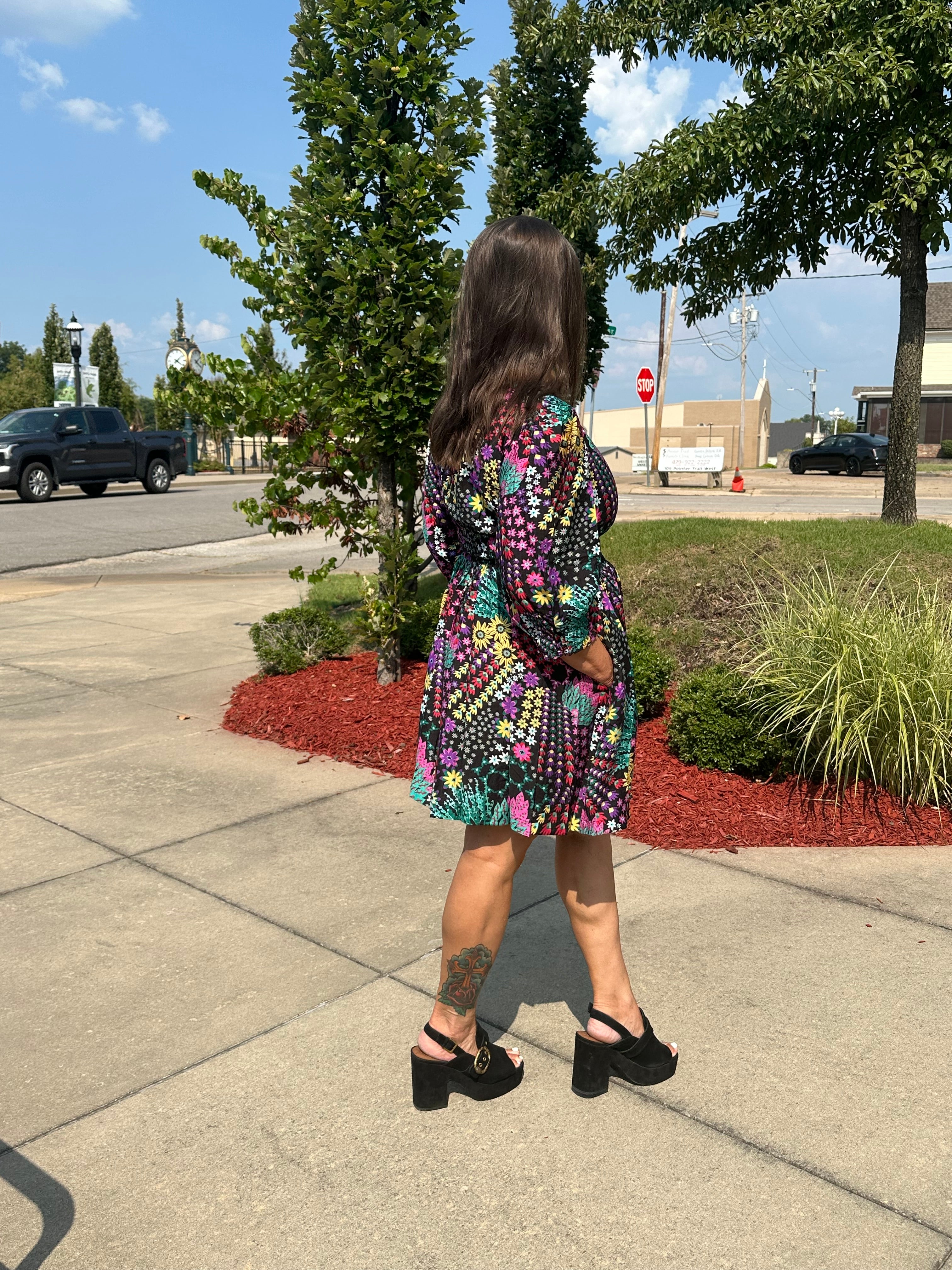 A woman is shown from behind wearing a colorful, short-sleeved patterned dress and black platform sandals while standing on a sidewalk outdoors.