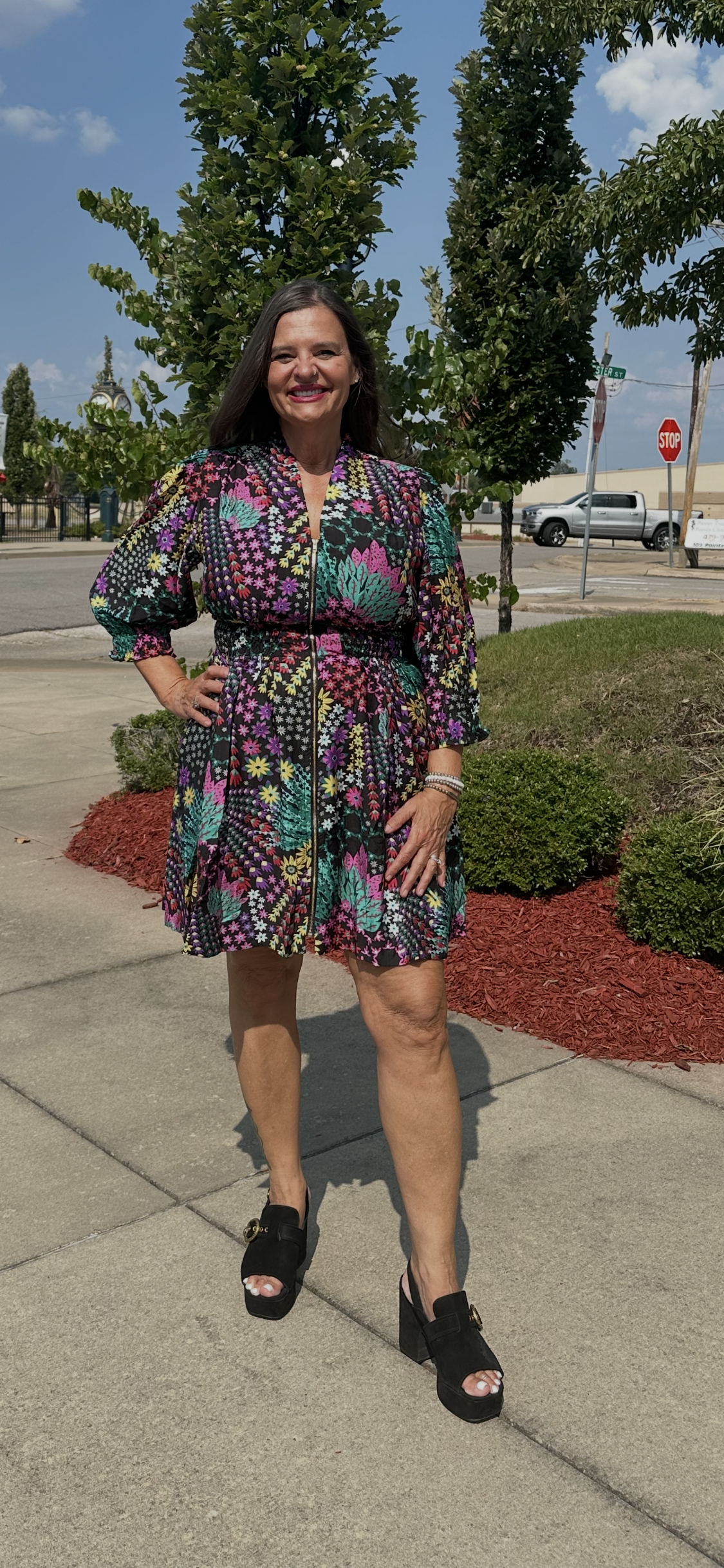 A woman wearing a colorful patterned mini dress with puff sleeves and black wedge sandals, standing on a sidewalk outdoors in front of trees and landscaping.