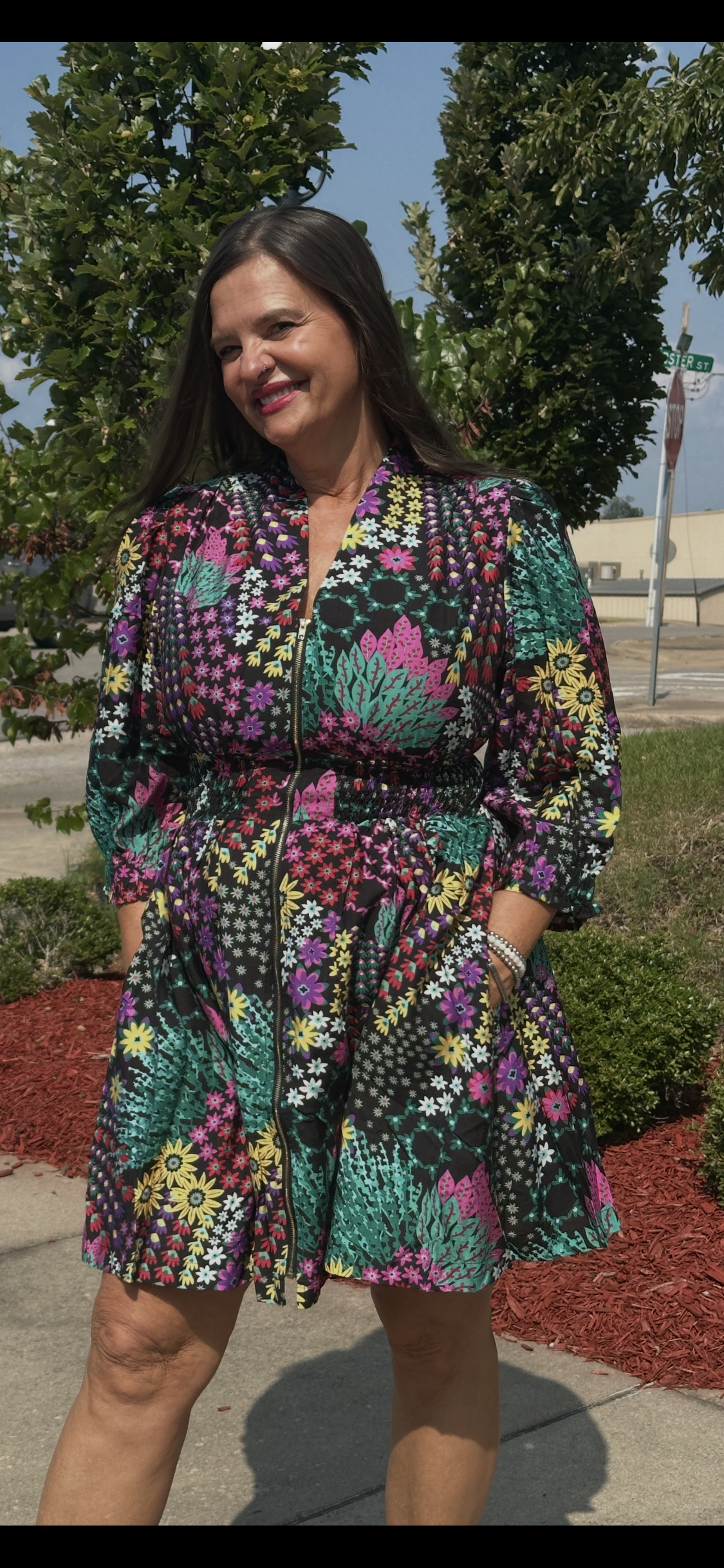 A woman wearing a colorful floral print long-sleeve mini dress, standing outdoors on a sidewalk with trees and landscaping in the background.