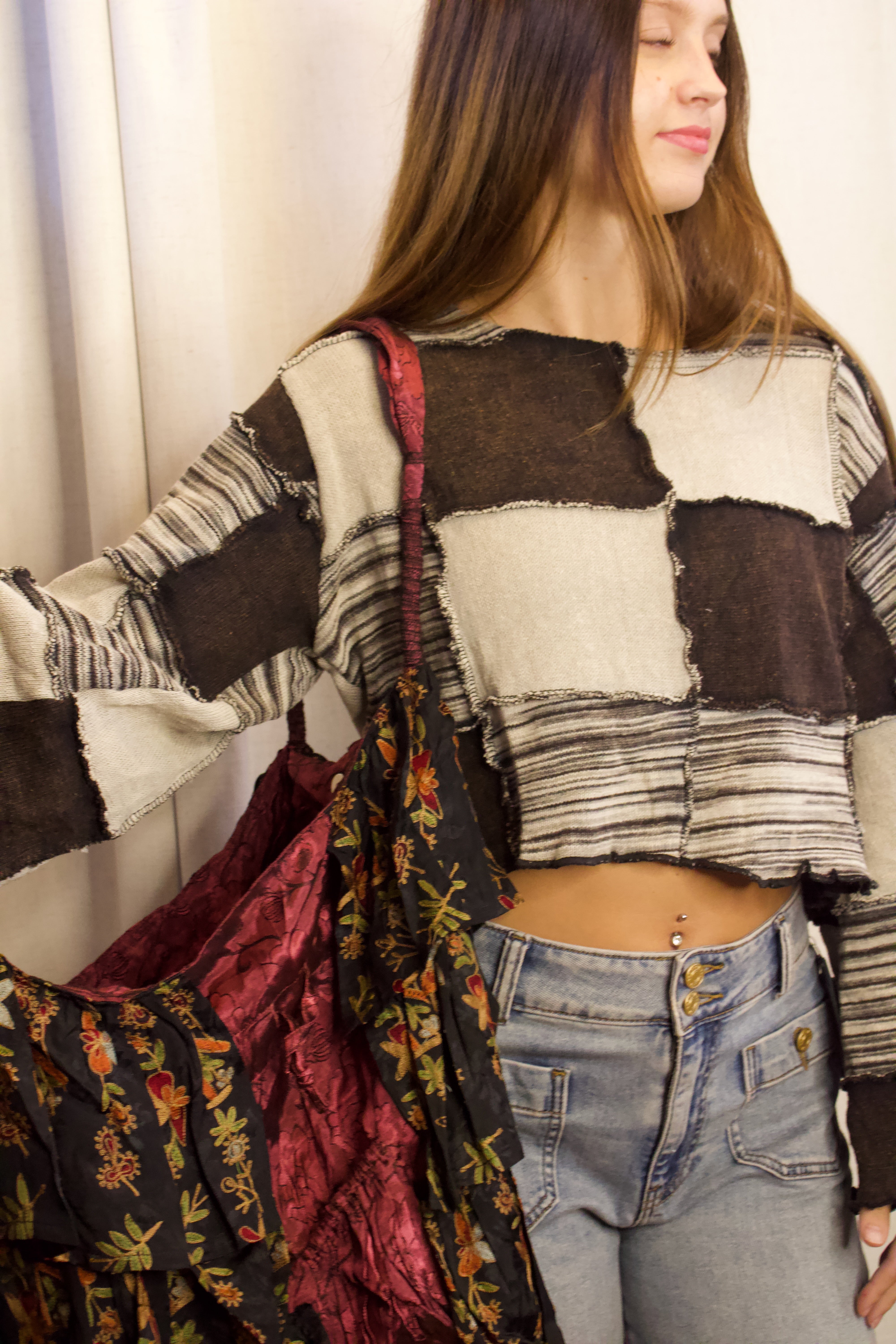 A woman wearing a patchwork long-sleeve crop top with brown, beige, and gray blocks, paired with light-wash jeans and carrying a large dark floral tote bag.